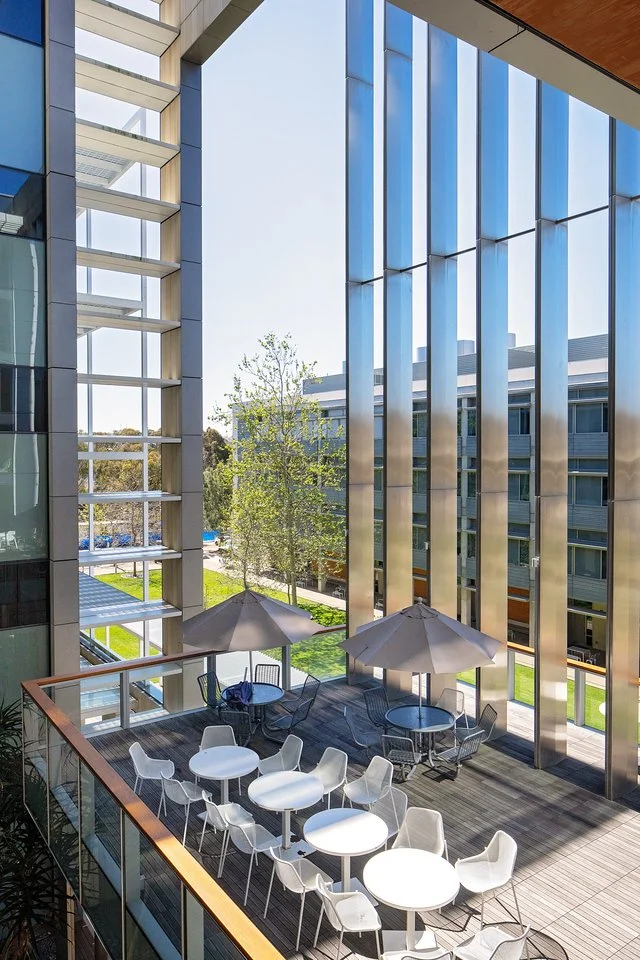 An outdoor patio with round tables, white chairs, and umbrellas surrounded by modern office buildings with large glass windows, green grass, and trees.