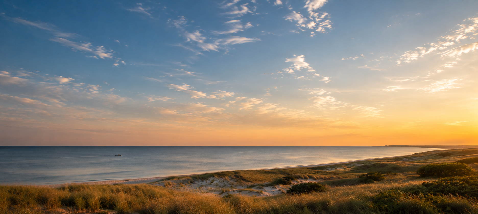 Sunset over a calm ocean with a sandy beach and grassy dunes in the foreground.