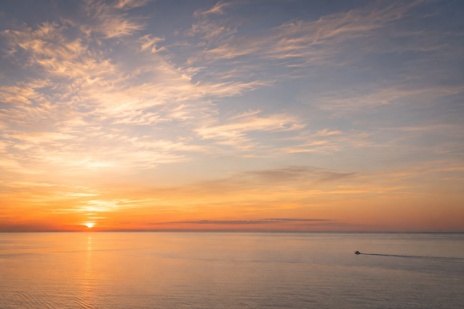 Sunset over calm ocean with a small boat leaving a wake in the water.