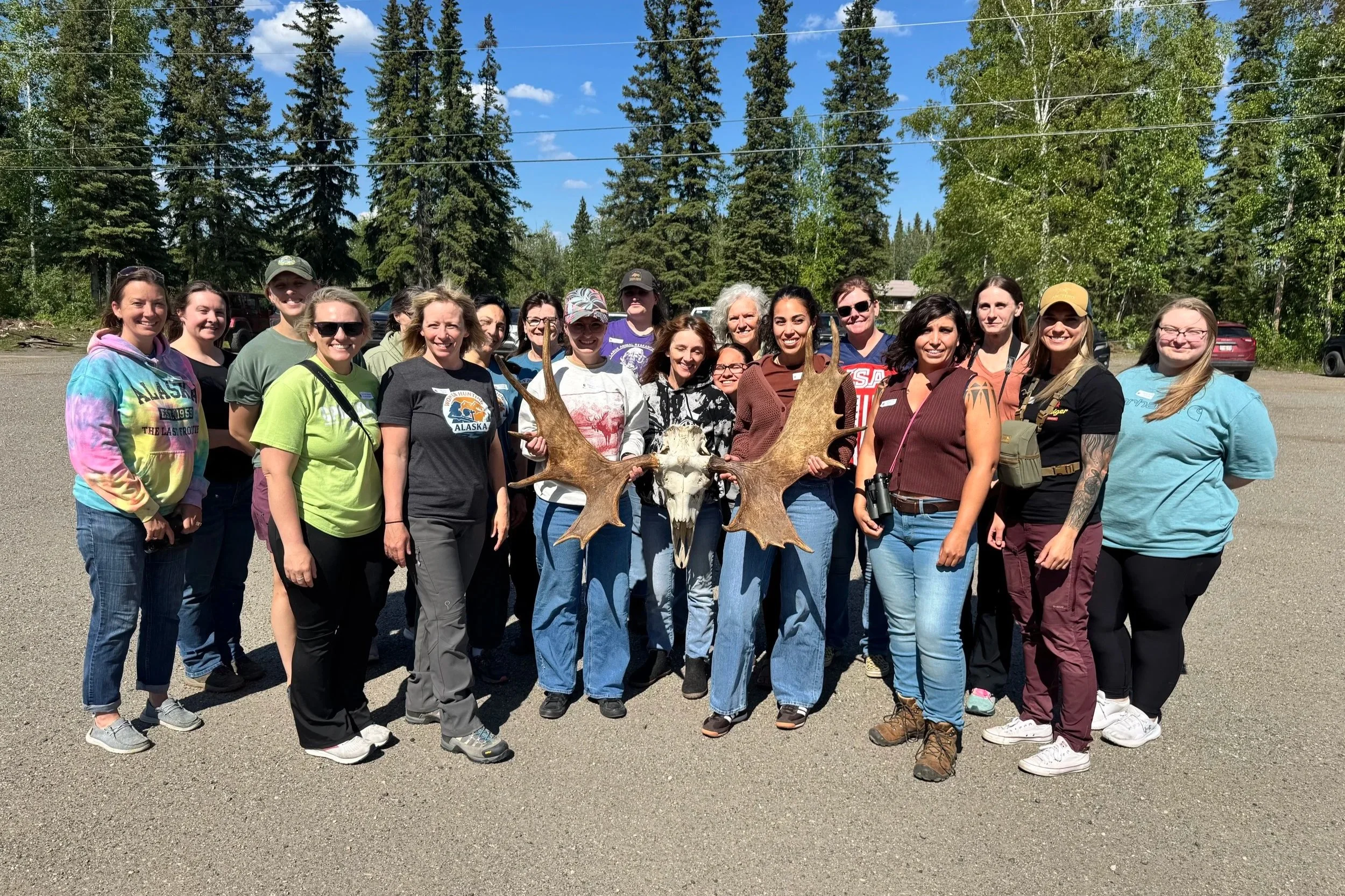 A group of people standing outdoors on a sunny day, holding a large moose skull with antlers. The background shows tall pine trees and a clear blue sky.