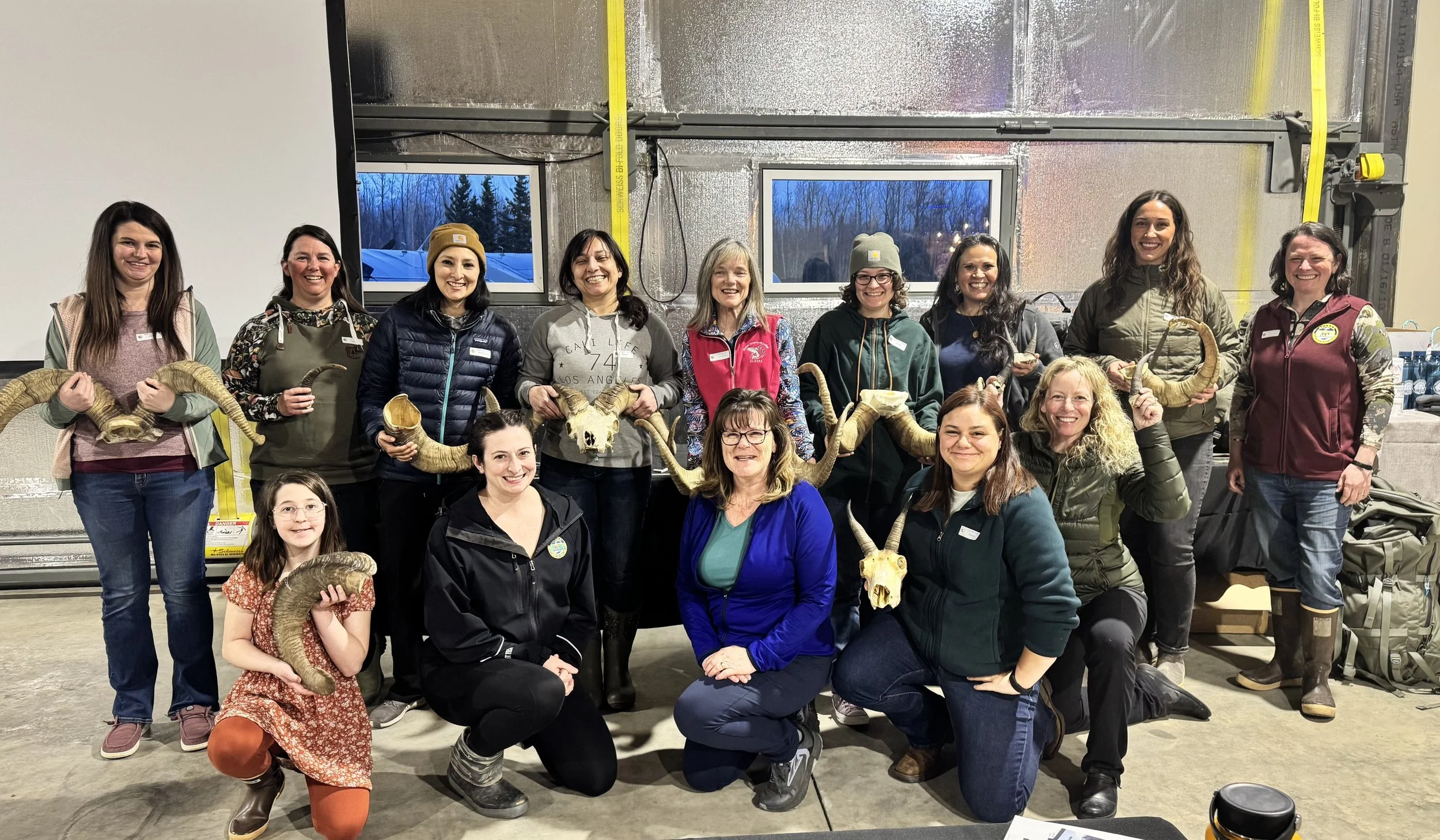 Group of women and children holding animal skulls and horns inside a garage with windows showing an outdoor winter scene.