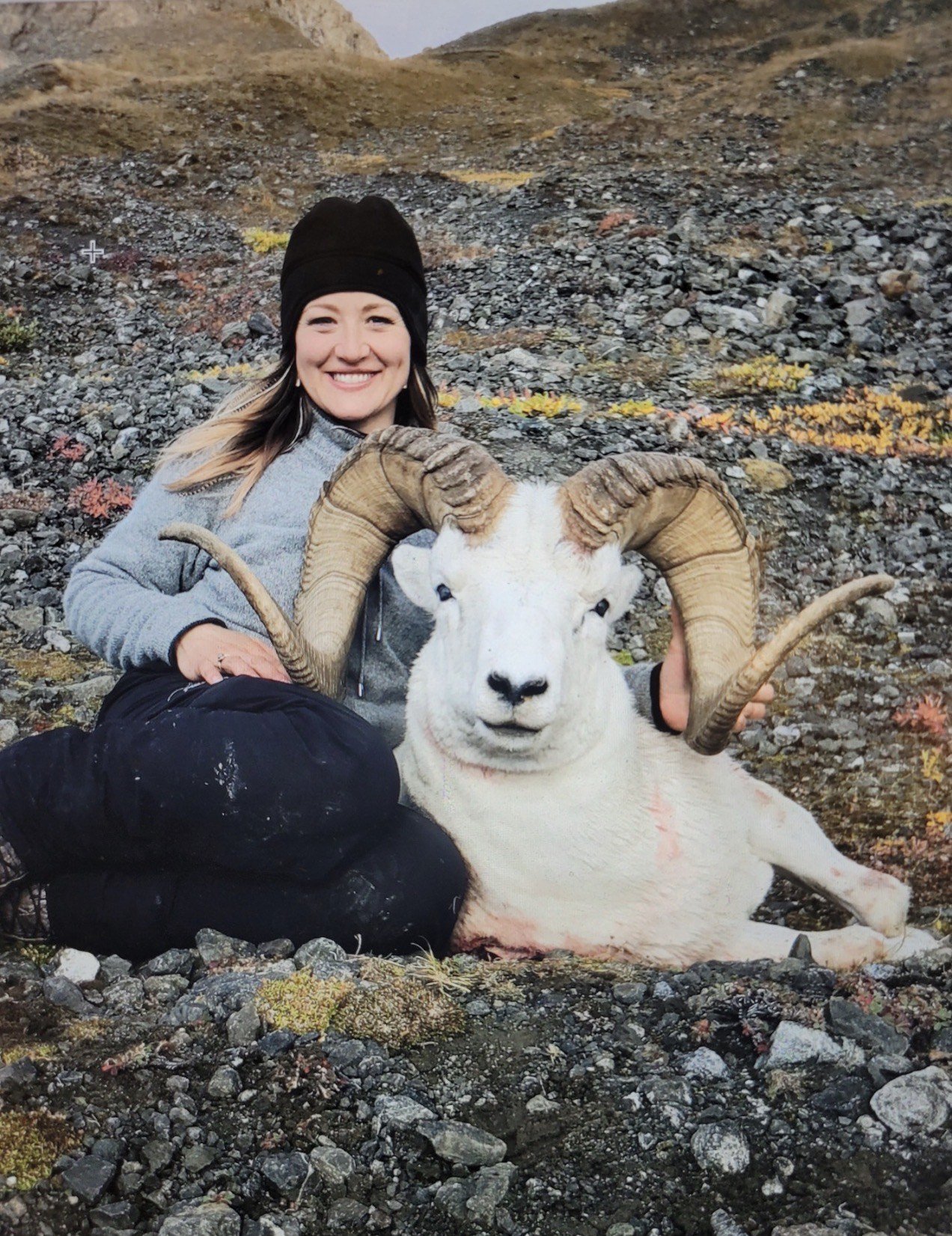 A woman sitting on rocky ground smiling and holding a large goat with prominent curved horns, in an outdoor mountainous area with sparse vegetation.
