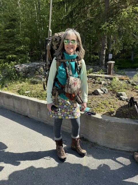 A smiling woman with gray hair, wearing sunglasses, a teal jacket, and brown hiking boots, stands on the side of a road with a large backpack and a walking stick in hand, in front of a wooded area.