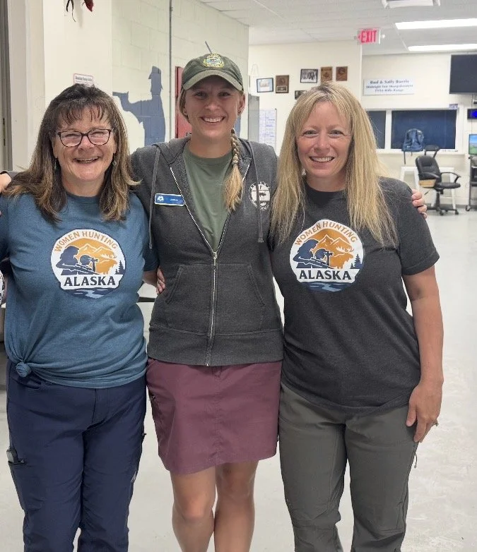 Three women standing indoors, smiling, with arms around each other, in an office or community center setting. The woman on the left and the woman on the right are wearing shirts with a logo that reads 'Women Hunting Alaska.'
