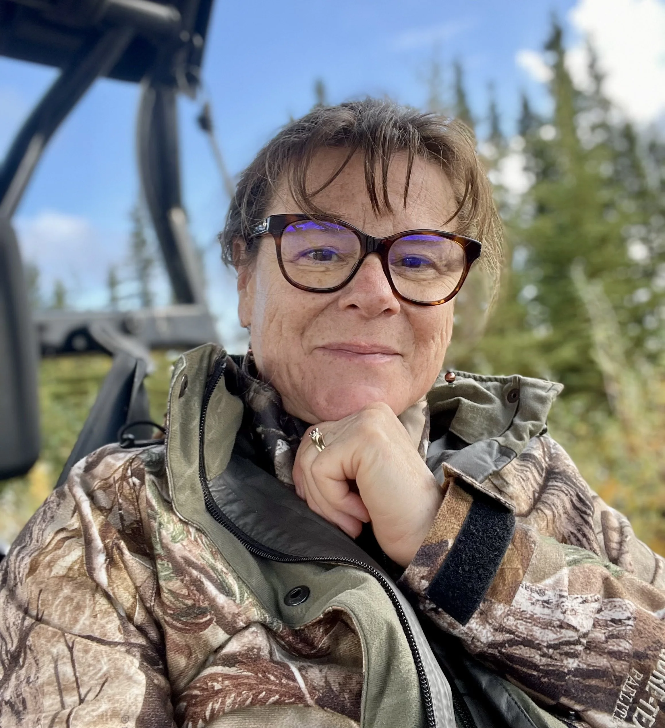 Close-up of an older woman with short brown hair wearing glasses and camo jacket, outdoors with trees and blue sky in the background.