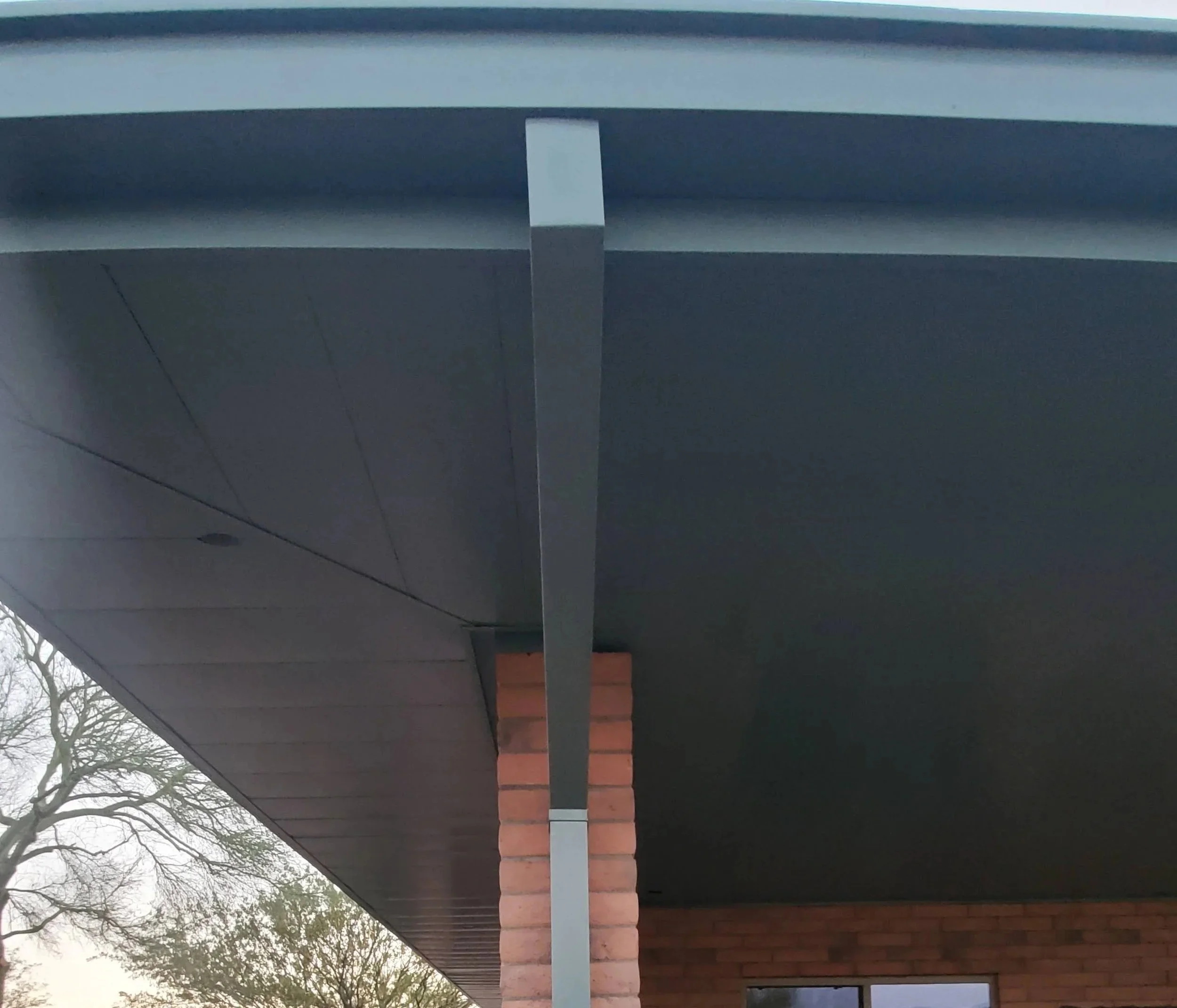 A close-up view of a wall with horizontal panels made of gray material and a section of red bricks, with the top edge featuring a white strip. Trees with bare branches are visible in the background under a cloudy sky.