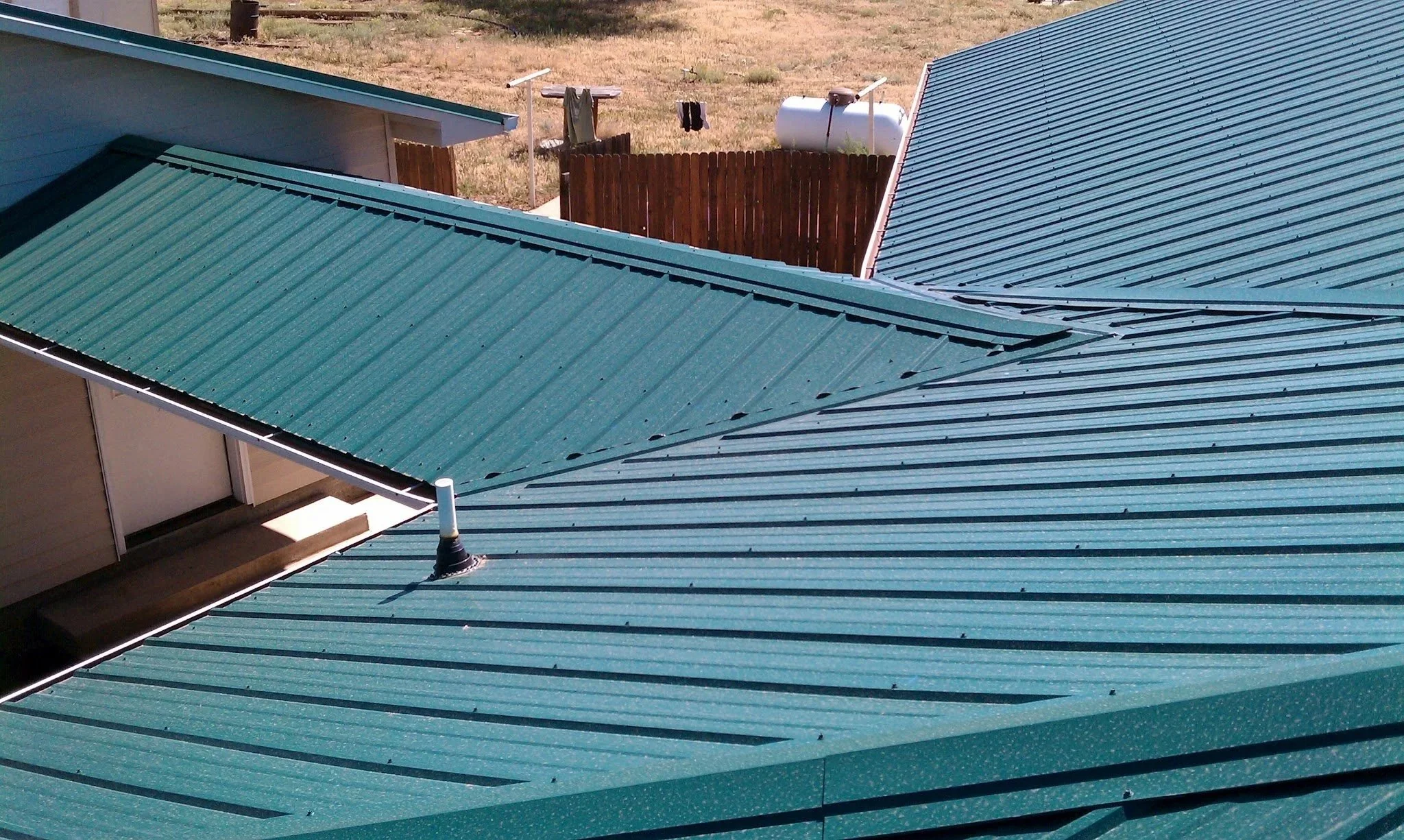 View of multiple green metal roofs with a fenced backyard area in the background, including a clothesline with hanging clothes, a cylindrical water tank, and a dry, brown lawn.