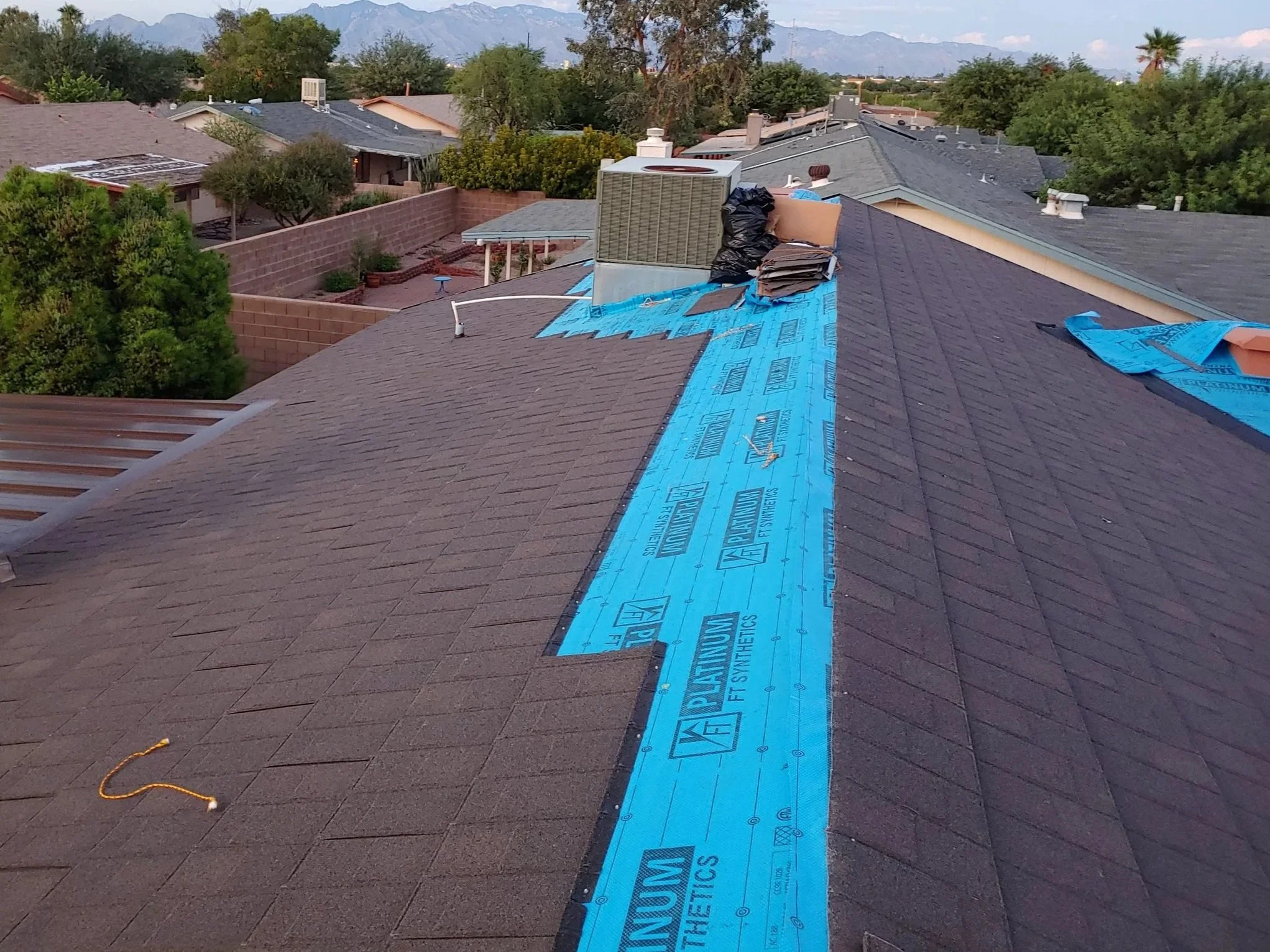 View of residential rooftops with construction materials and equipment on a roof, construction work in progress, blue insulation sheets, and a yard with trees and a brick wall, mountains in the background.