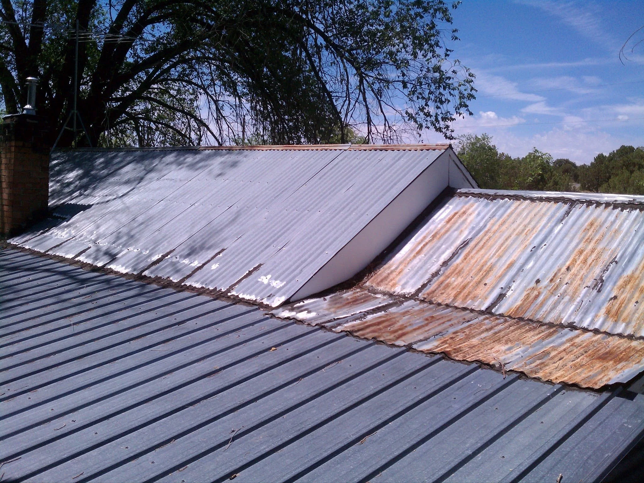Rooftop with corrugated metal sheets, some rusty and weathered, under a partly cloudy blue sky with trees in the background.