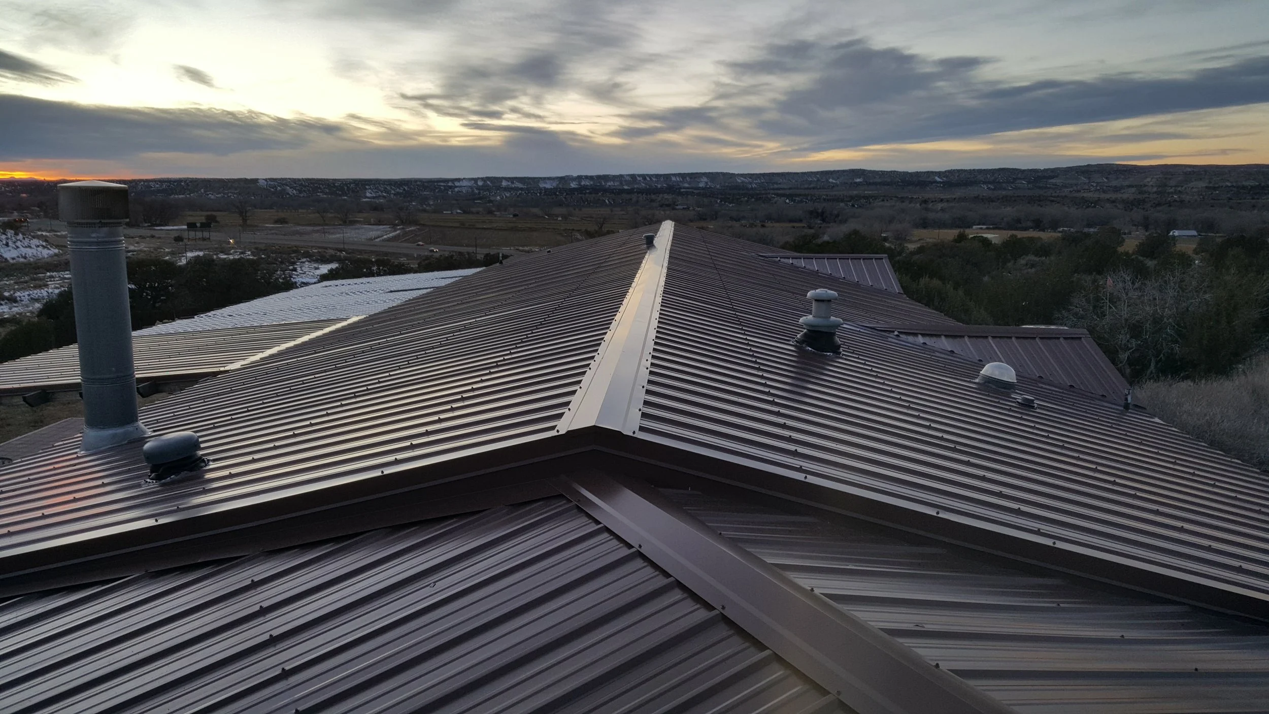 Metal roof with vents and a chimney, overlooking a landscape with hills, trees, and a cloudy sky at sunset.