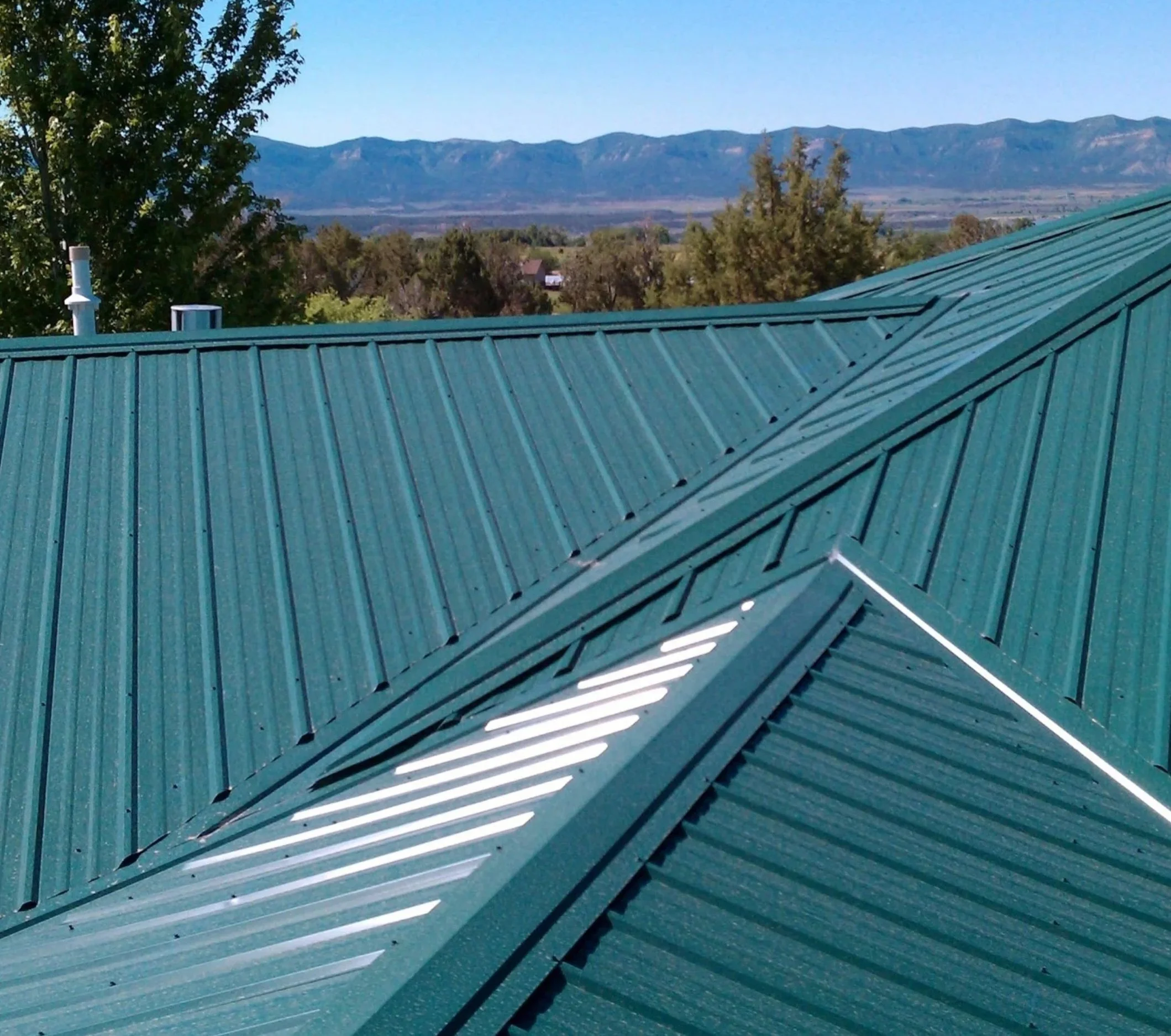 A teal-colored metal roof with ridges, overlooking a landscape of trees and a mountain range in the background under a clear blue sky.