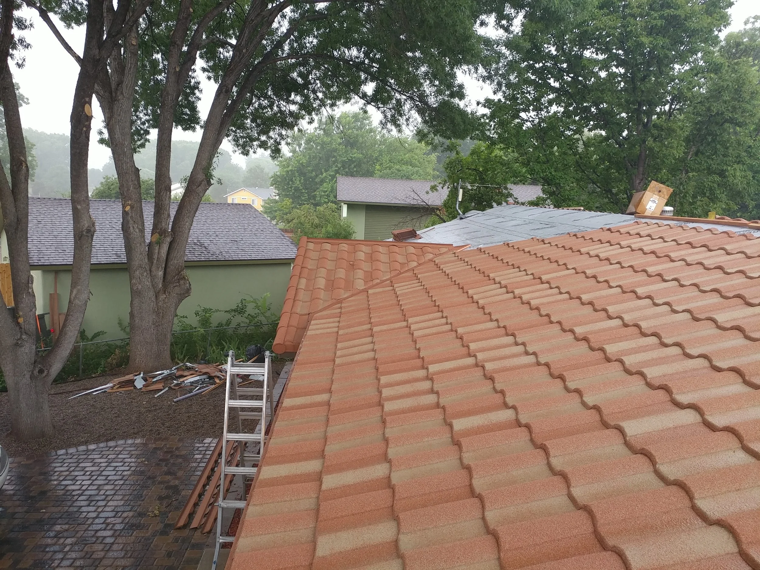 A view of several houses with tile roofs, some under construction or repair, in a suburban neighborhood surrounded by tall green trees and overcast weather.