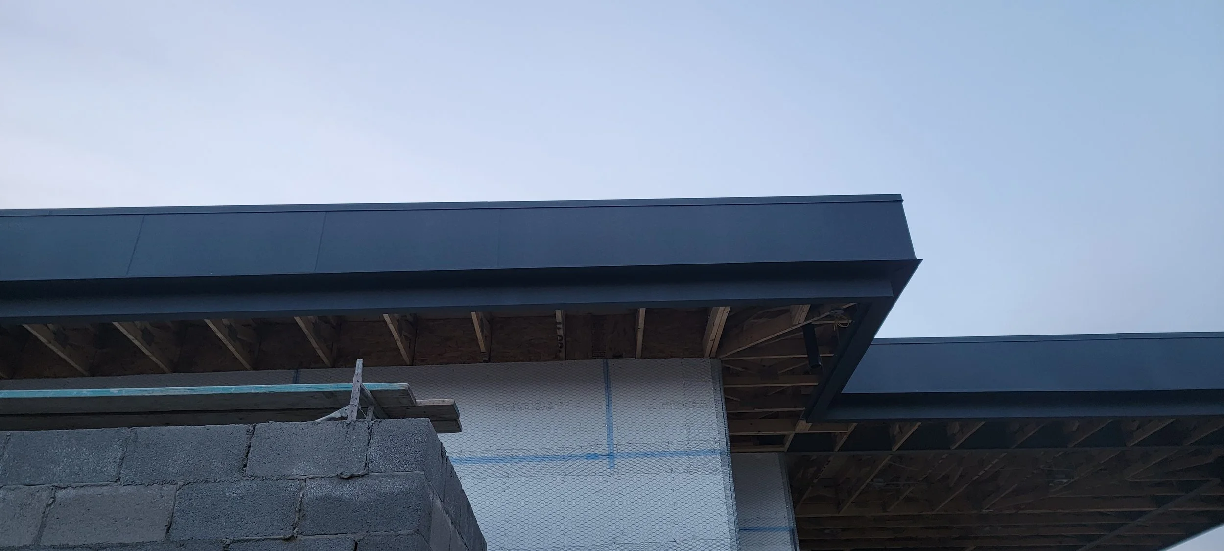 Part of a building under construction with exposed wooden beams, gray siding, and a brick wall in the foreground against a clear sky.