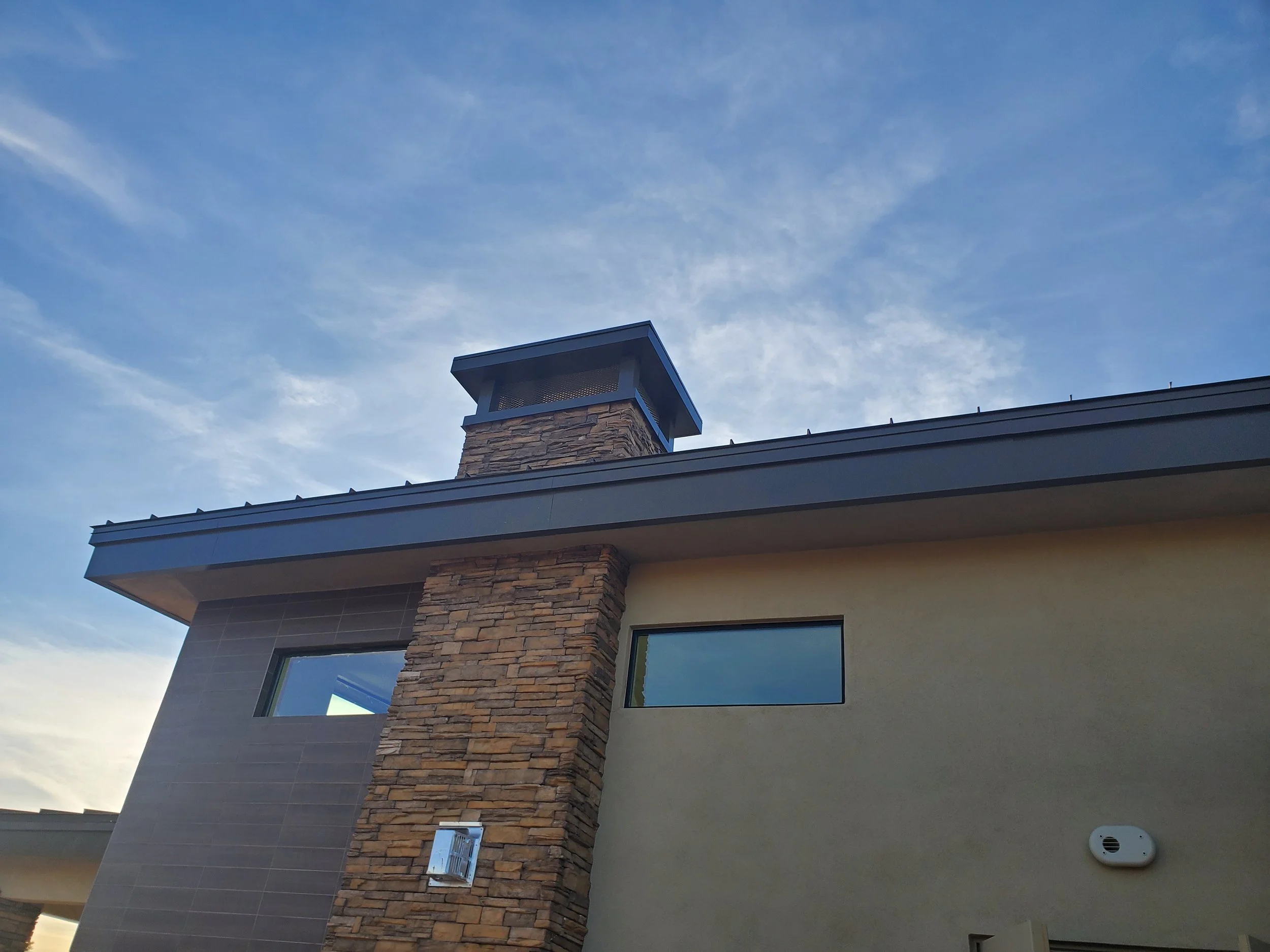 Close-up view of the corner of a building with a chimney made of brick and a vent on the wall under a blue sky with some clouds.