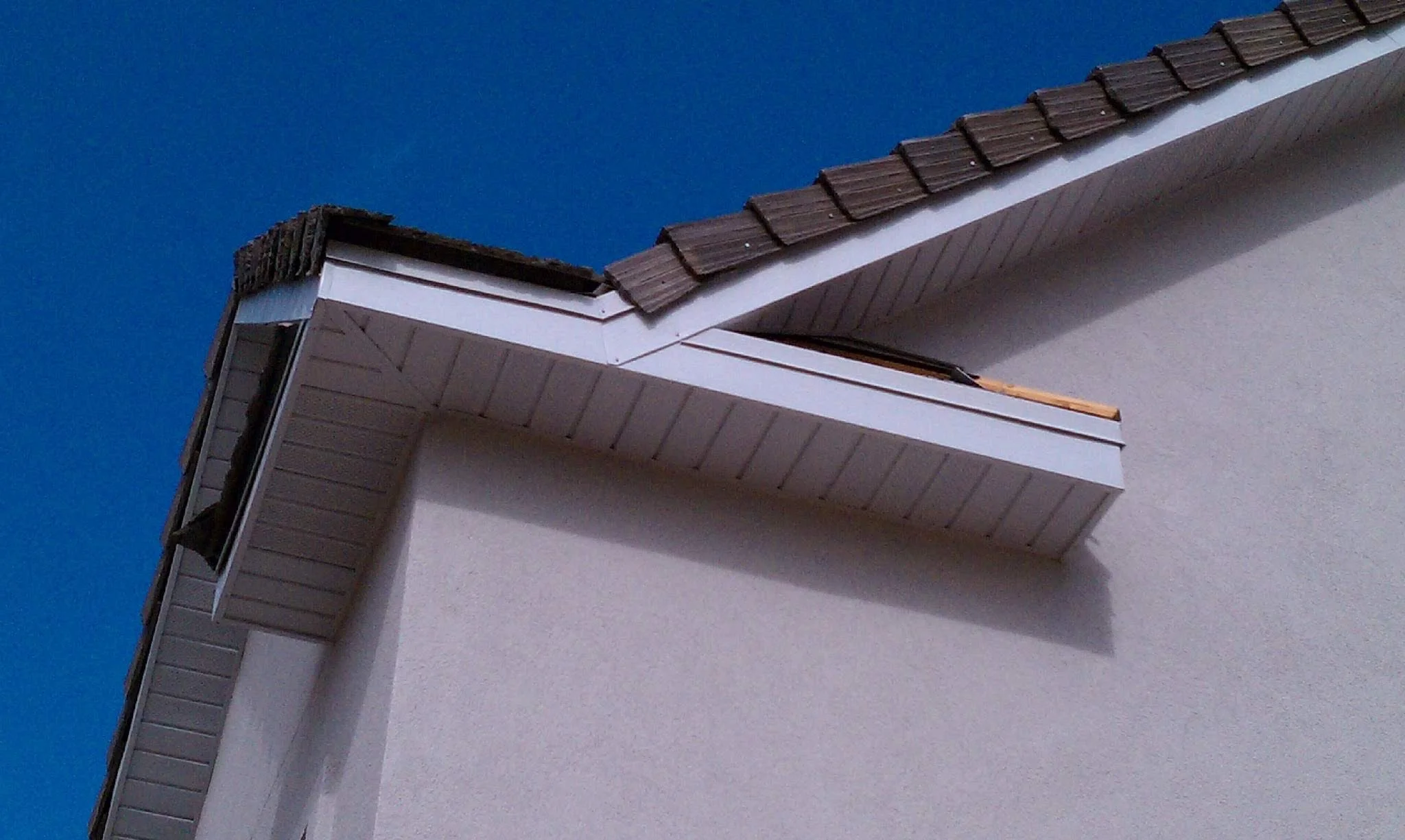 Close-up of a house corner with gray shingles on the roof and a white gutter against a clear blue sky.
