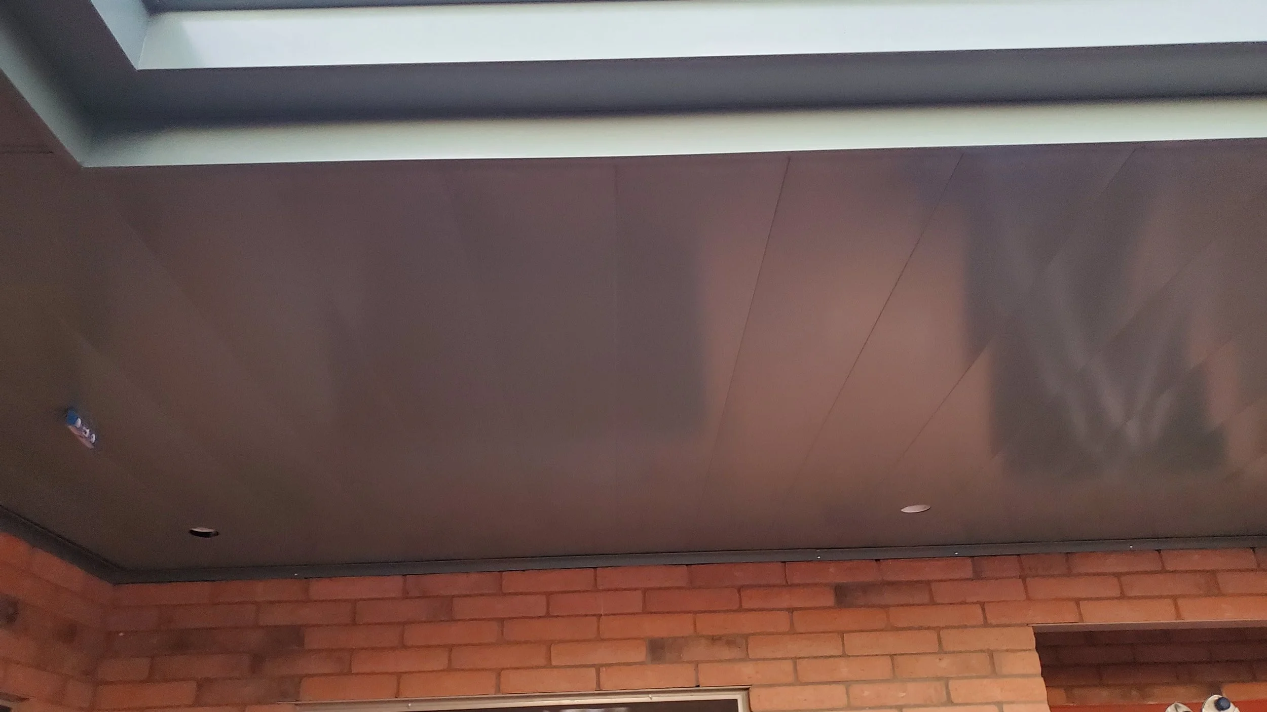 The image shows a ceiling mounted metal ventilation or exhaust hood with a dark grey surface, placed against a brick wall. There are small visible holes in the hood, and a small part of a fire alarm or smoke detector can be seen in the corner.