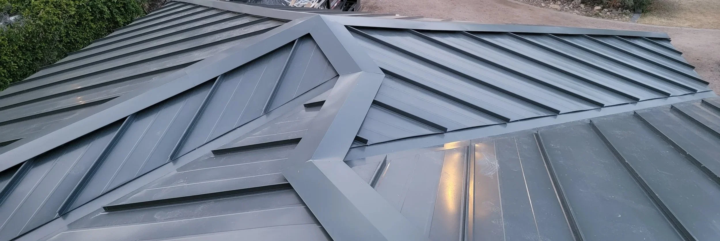 Close-up of a modern, gray metal roof with angular design and ridges, with greenery and dirt ground in the background.