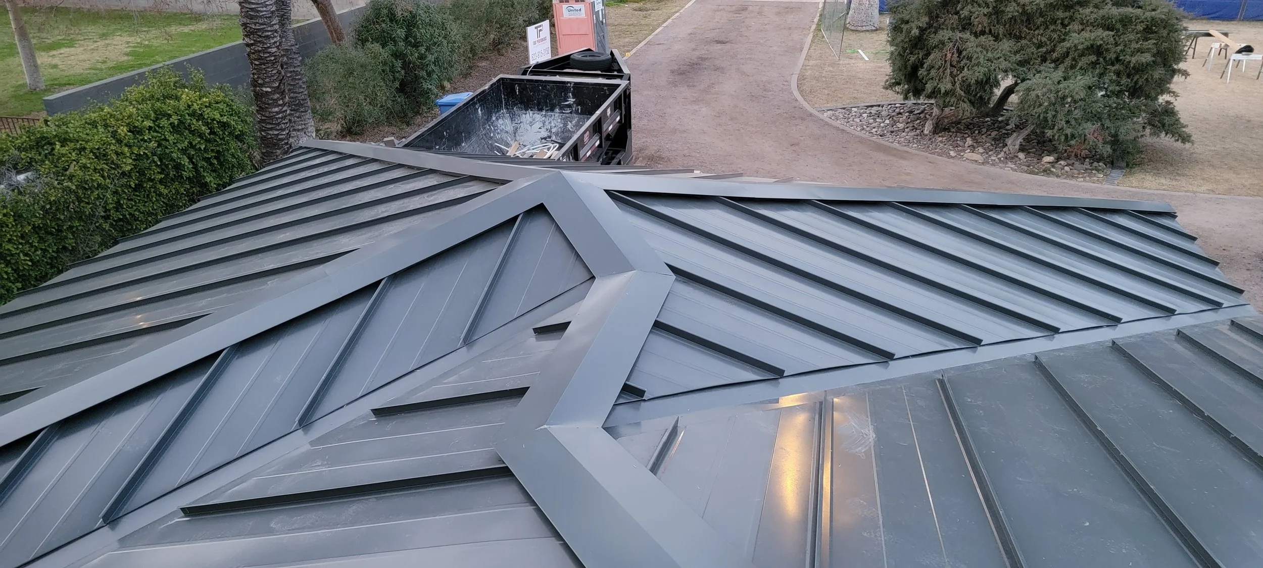 Metal rooftop with a solar panel installed on it, surrounded by trees and a dirt pathway.