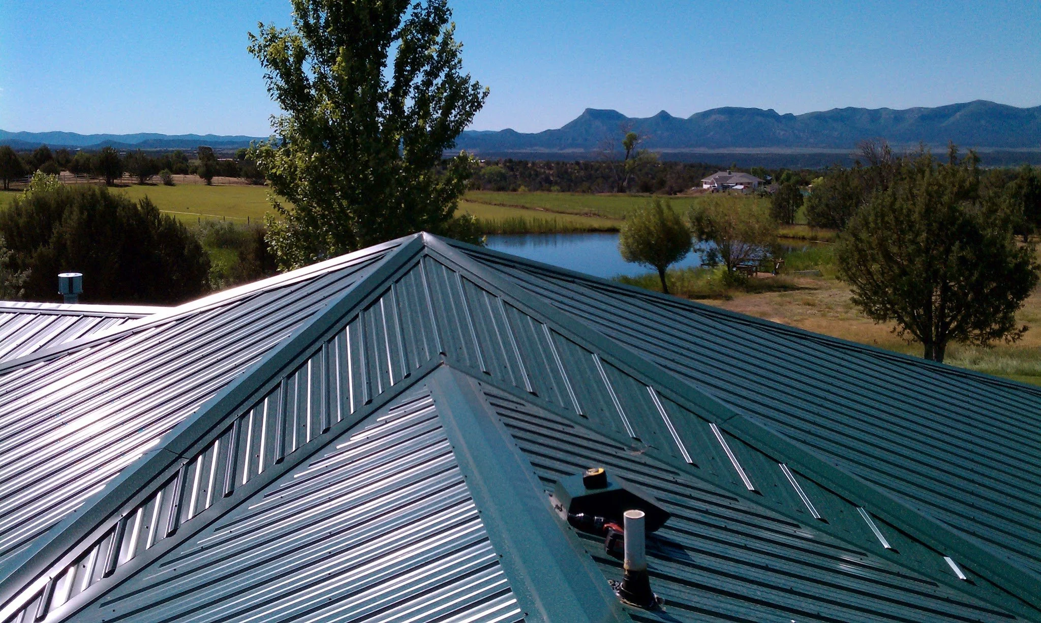 Metal roof with skylights and vent pipes, overlooking a rural landscape with trees, water, grass, and mountains in the distance.