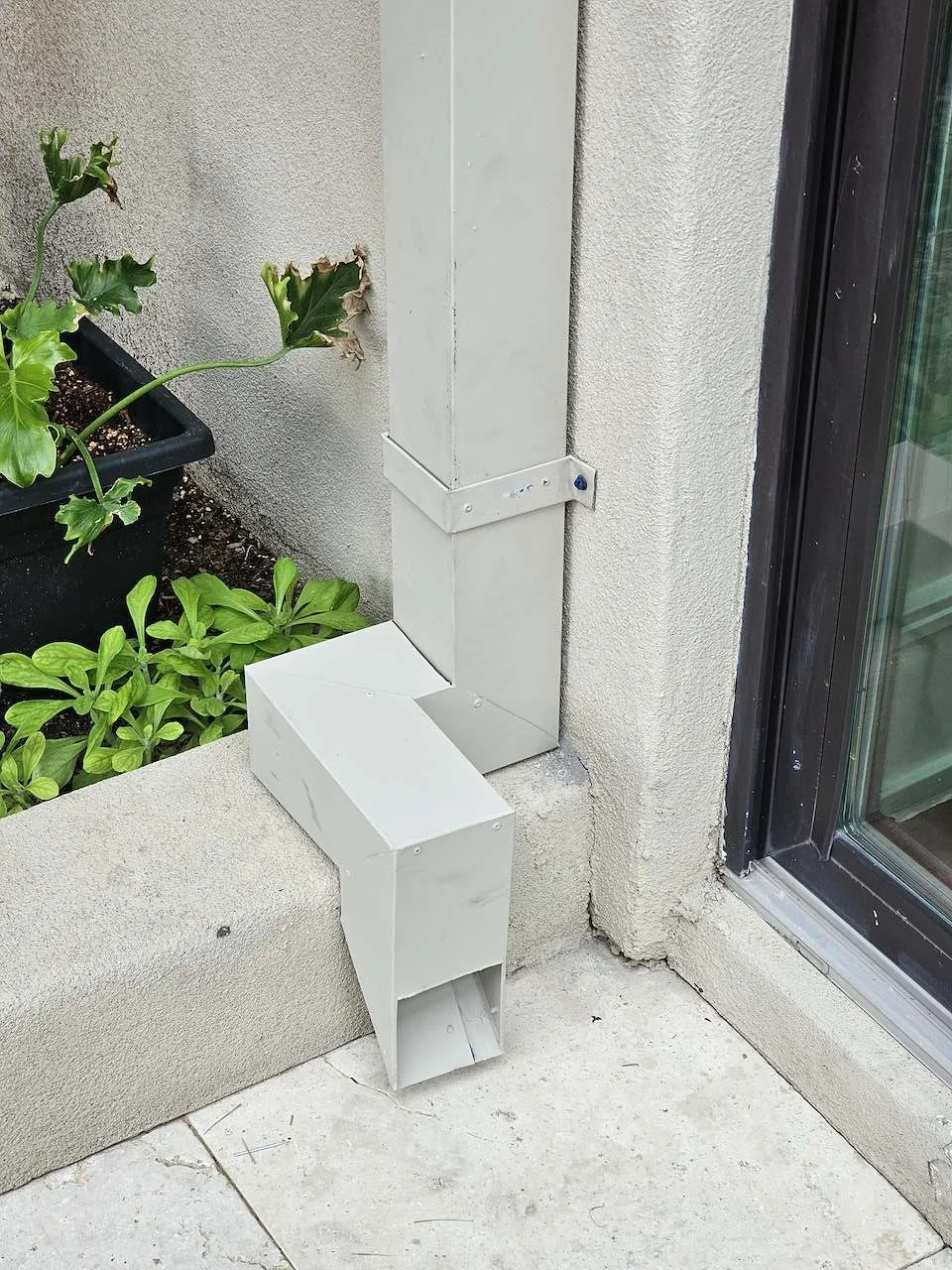 Close-up of a metal downspout and electrical conduit on an exterior wall near a sliding glass door, with plants nearby.