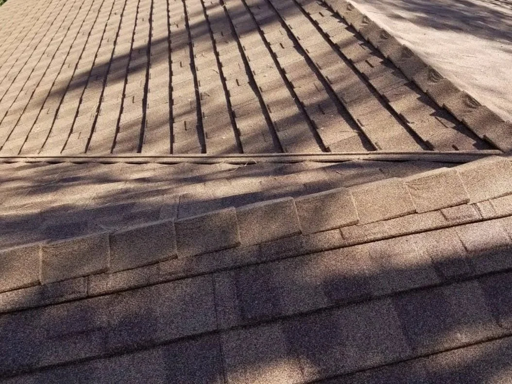 Close-up of a sloped roof with asphalt shingles and a section of wooden roof tiles in sunlight, with shadows cast across the surface.