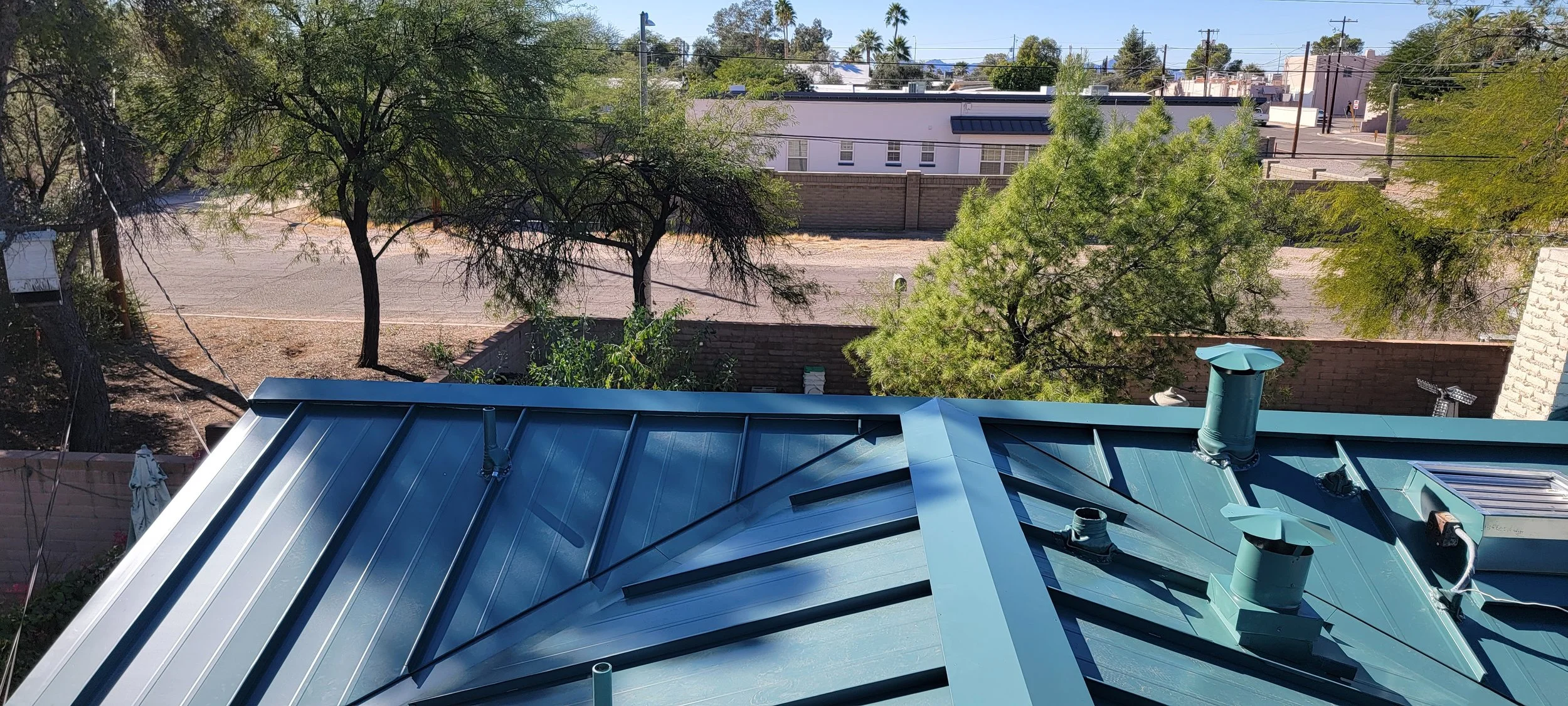 View from a rooftop showing a metal roof with vents, a yard with trees, a street with parked cars, and buildings in the background.