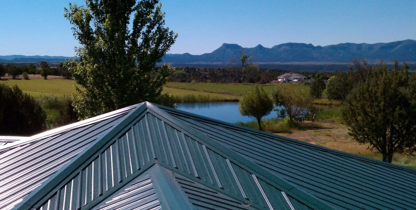 View of a metal roof with a lap seam design, overlooking a tranquil landscape featuring a pond, green trees, open fields, and distant mountains under a clear blue sky.