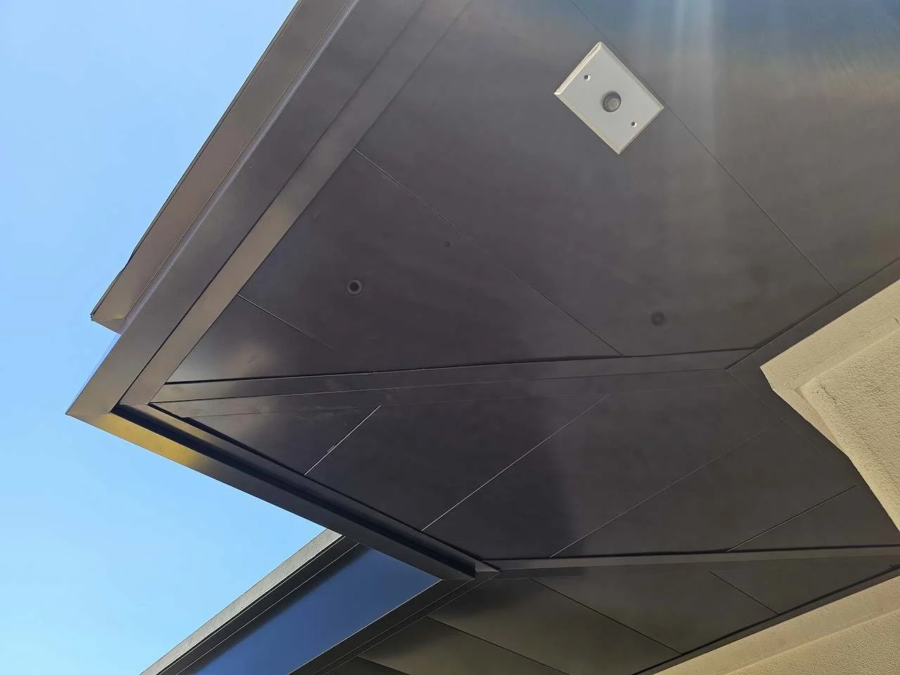 Close-up of a modern building's exterior corner featuring dark metal panels, a beige textured wall, and a blue sky background.