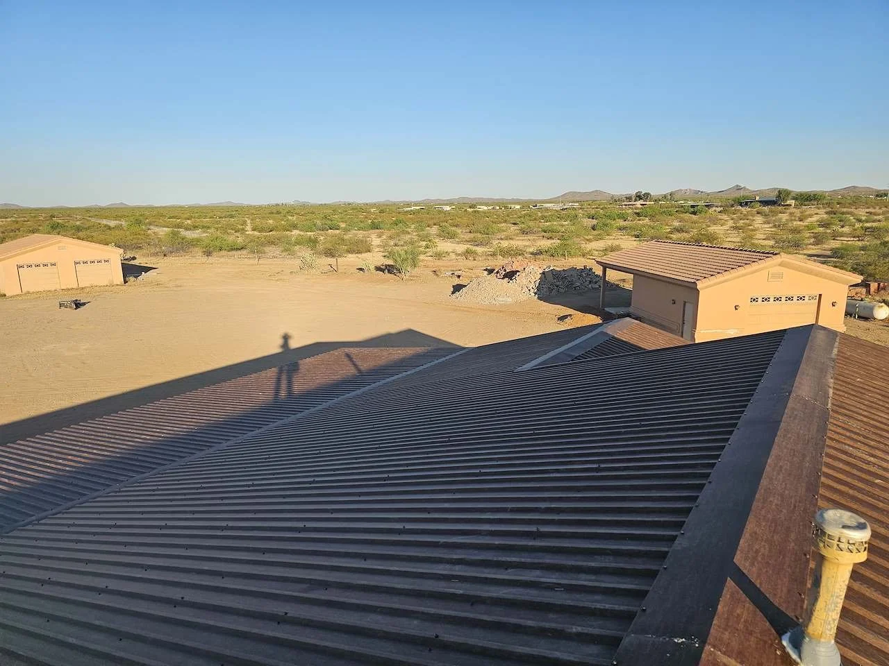 View of a dry, semi-arid landscape with distant mountains under a clear blue sky, featuring the roofs of two houses with metal and shingle roofing.