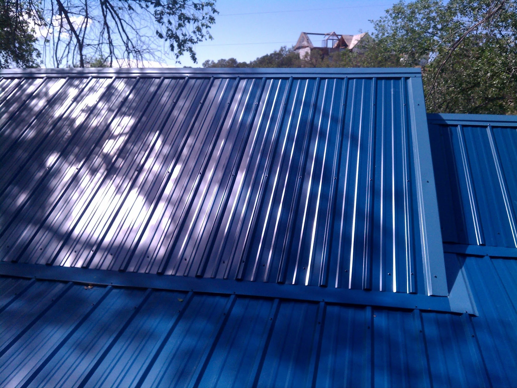 Photo of a blue metal roof with trees and a house in the background, under a clear blue sky.