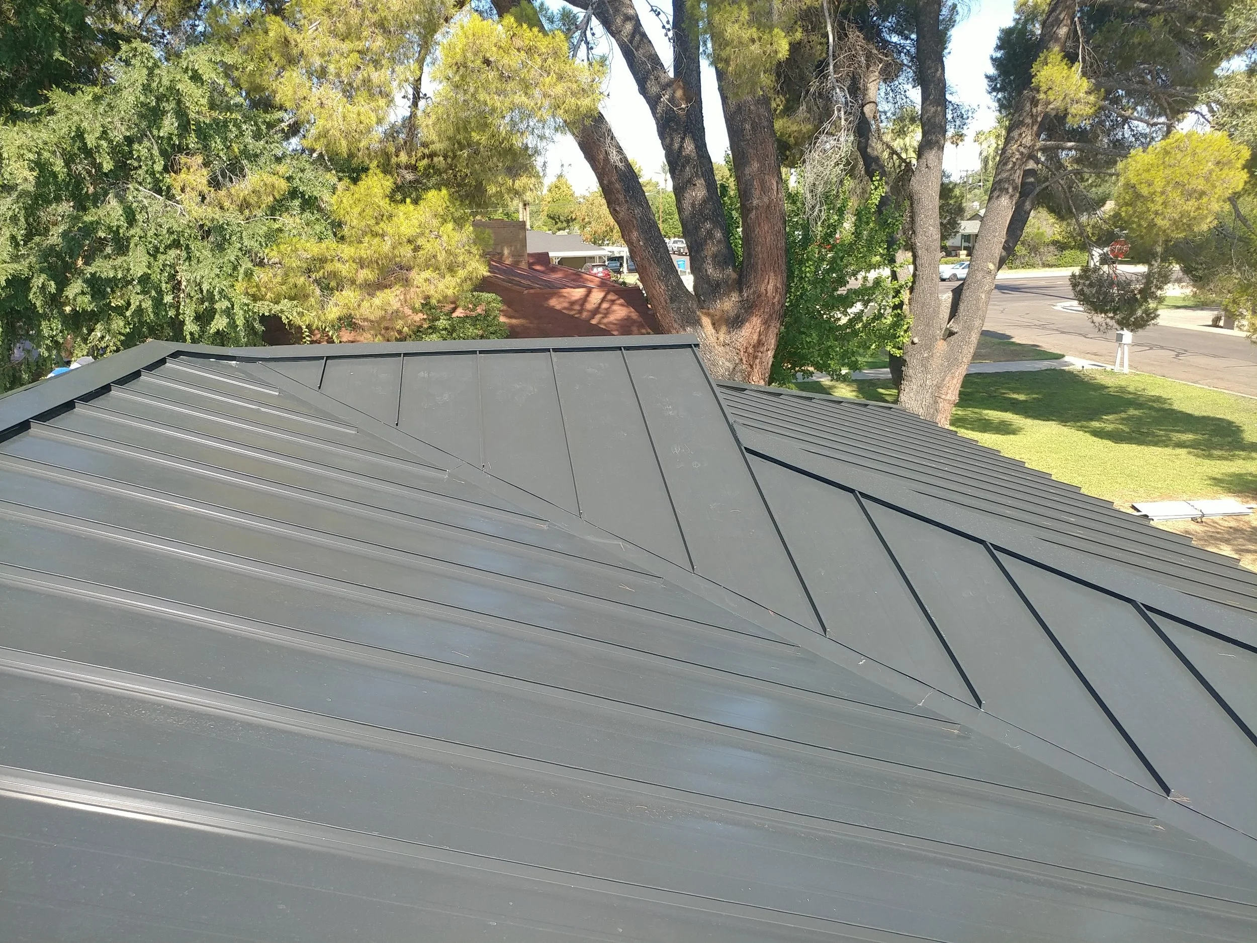 View of a gray metal roof in the foreground with green trees and a street with parked cars in the background.