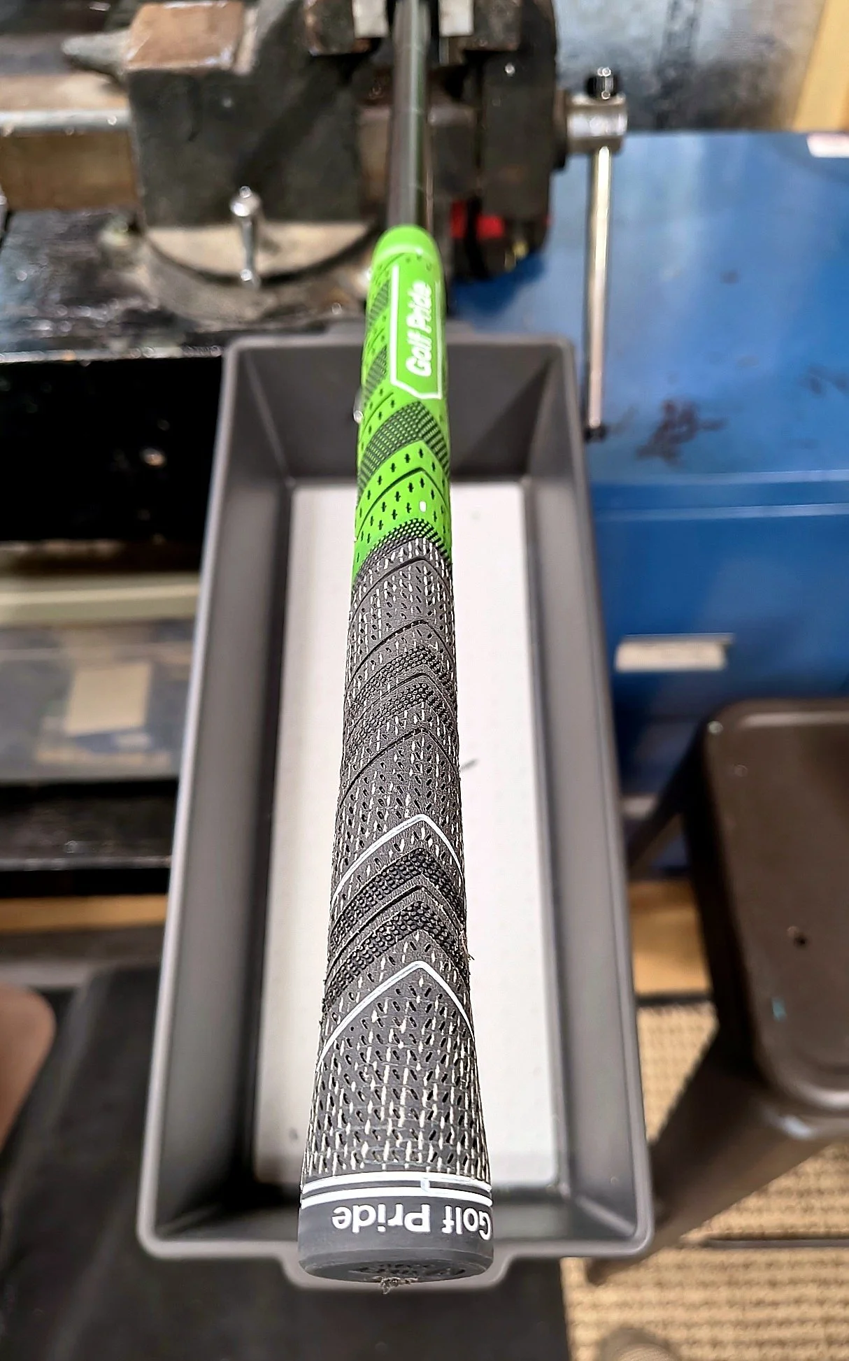 A golf putter with a grey grip, placed inside a grey golf club holder. The grip has intricate black and white designs with the words "GOLF Prite" printed at the bottom. The background includes a cluttered workbench with tools and equipment.