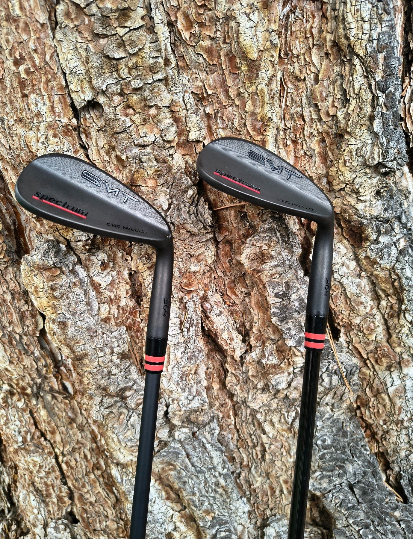 Two golf clubs with black grips leaning against tree bark.