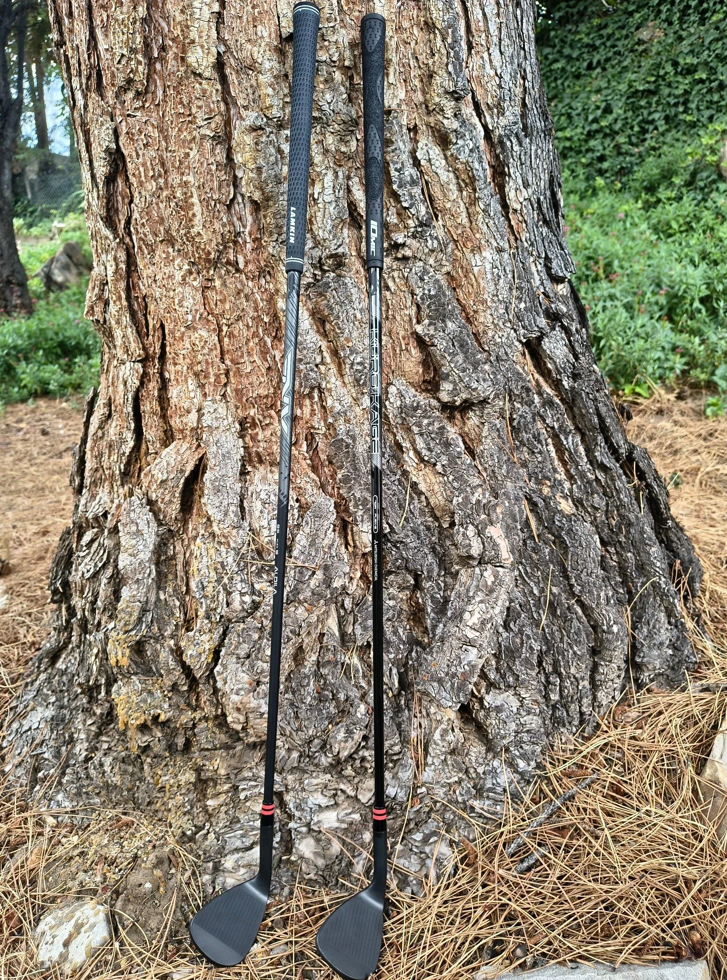 Two golf clubs with replacement grips leaning against a tree outdoors.