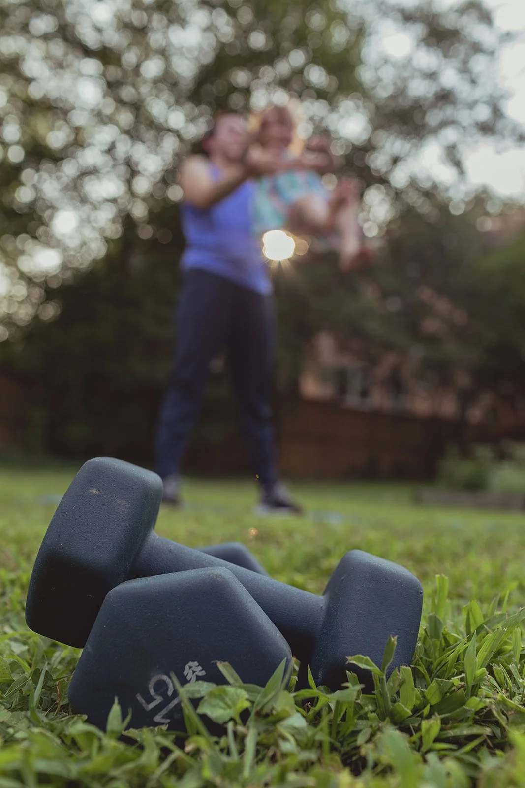 A pair of black dumbbells on grass in the foreground, with a man and a child out of focus in the background, seemingly playing or lifting a child outdoors during daylight.