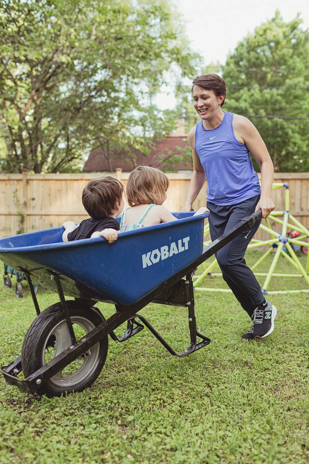 A woman pushing a blue wheelbarrow with two children sitting inside in a backyard with trees and a wooden fence.