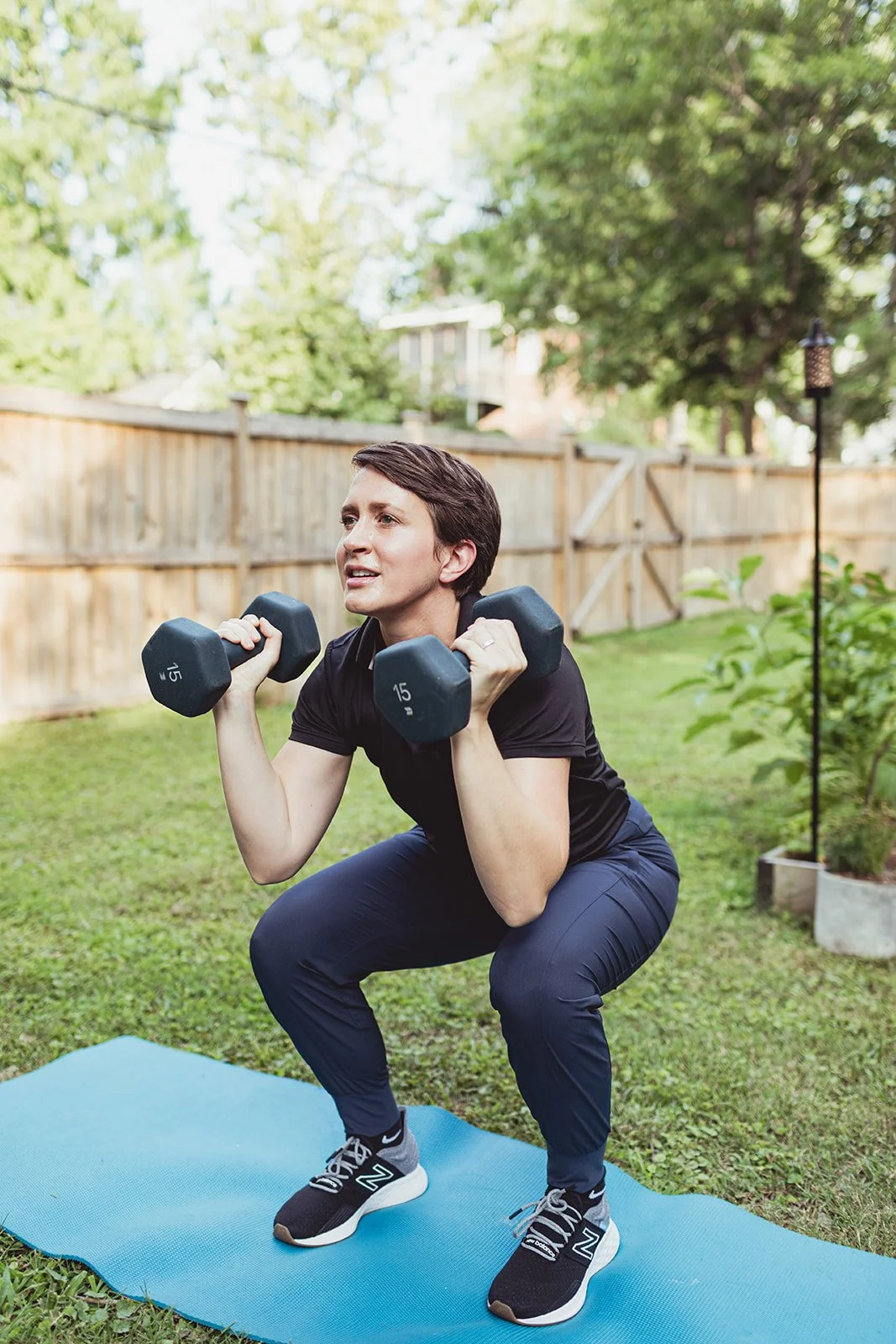 A woman exercising outdoors on a blue yoga mat, squatting while holding dumbbells near her shoulders, with a wooden fence and trees in the background.