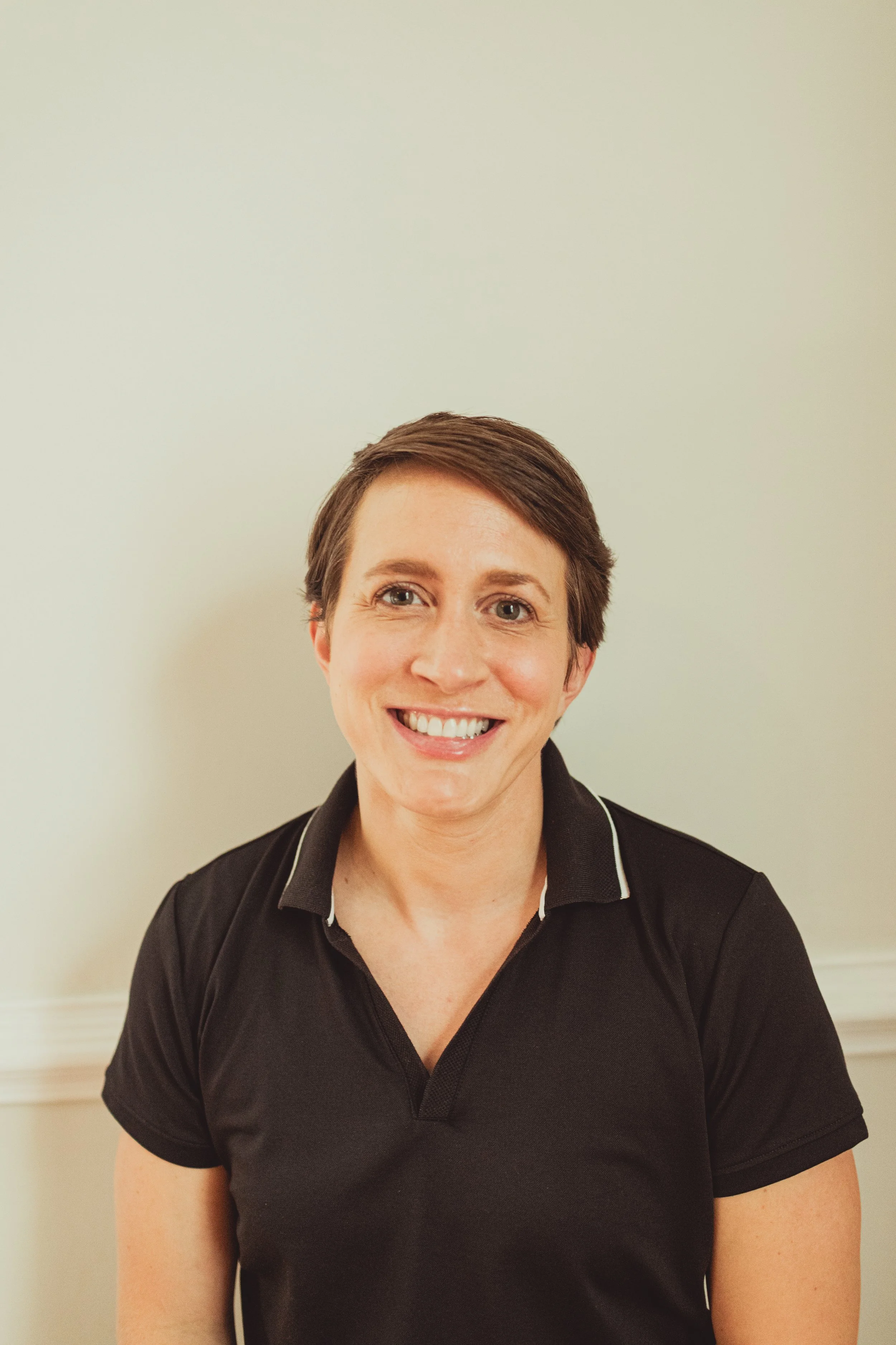 A woman with short brown hair and a big smile, wearing a black polo shirt, standing against a plain beige wall.