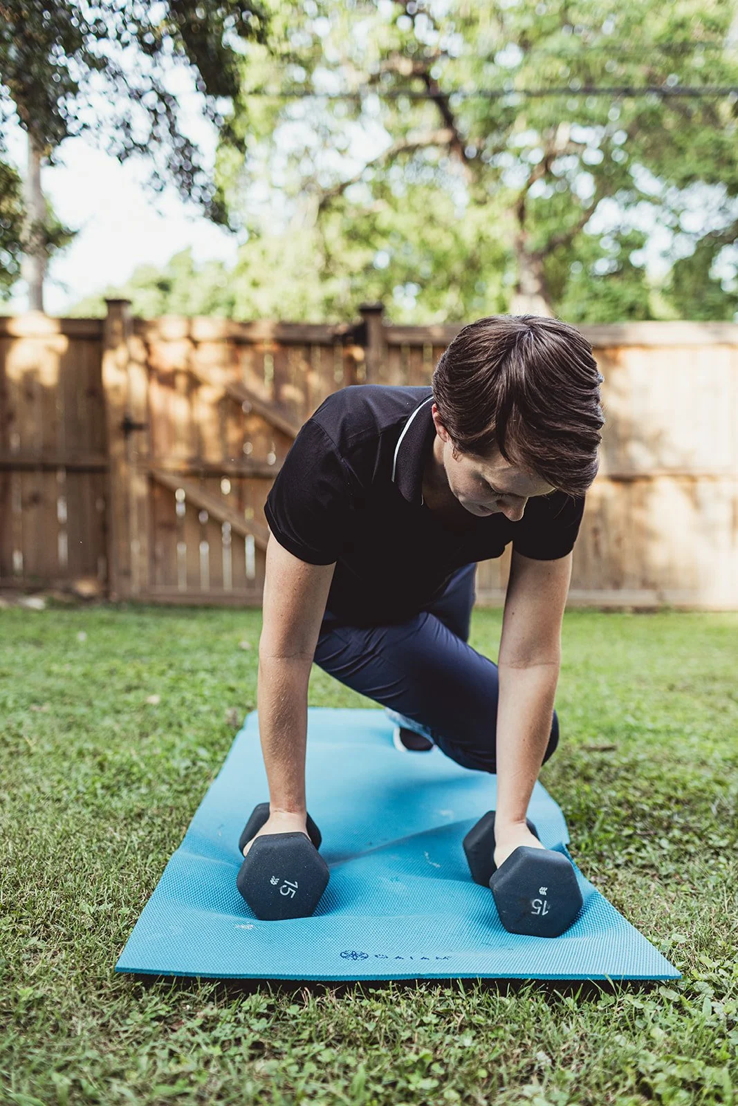 A woman exercising outdoors on a yoga mat, holding a dumbbell in each hand, with a wooden fence and trees in the background.