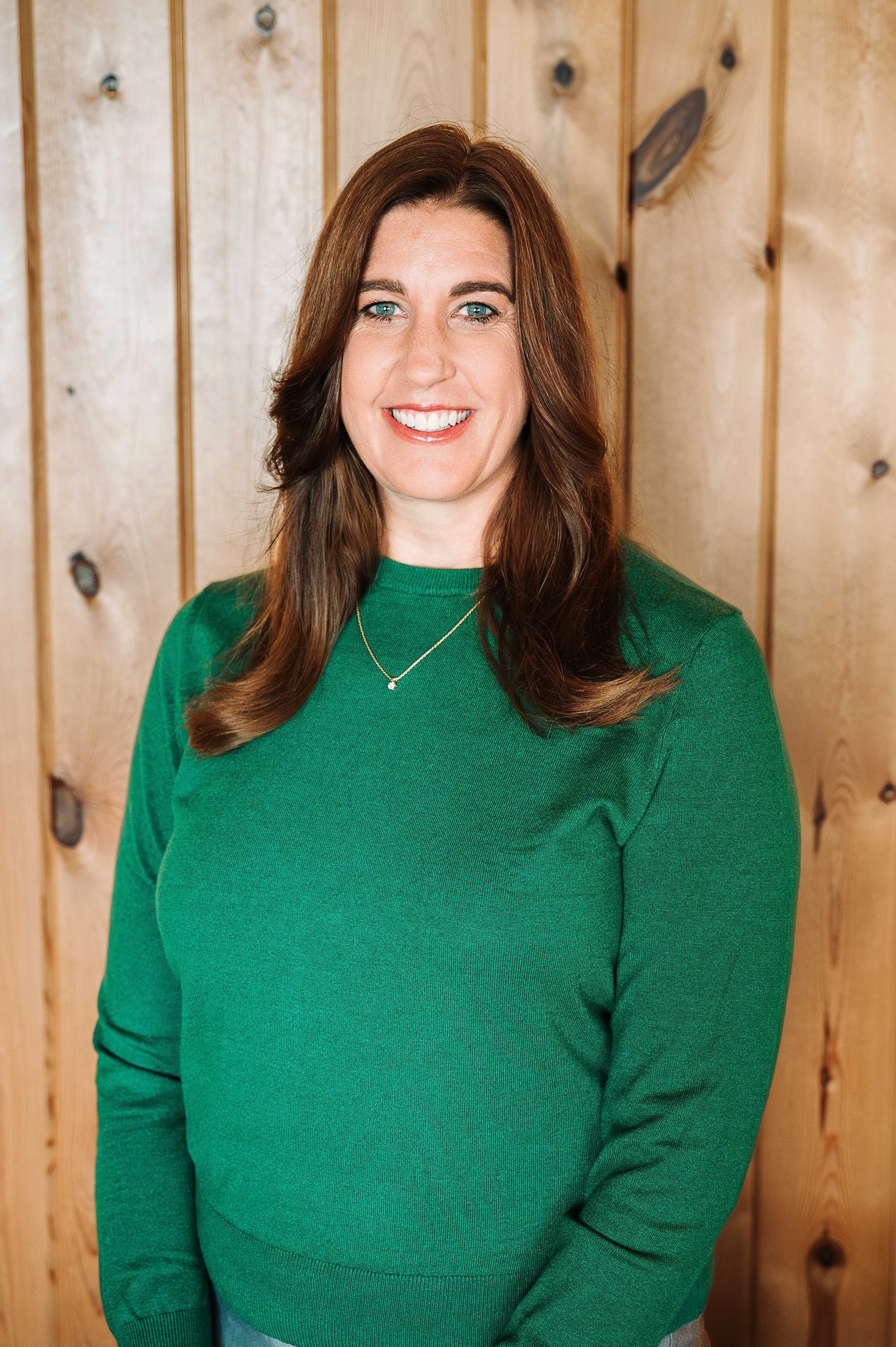 A woman with long brown hair and blue eyes smiling, wearing a green sweater and a silver necklace, standing in front of a wooden wall.