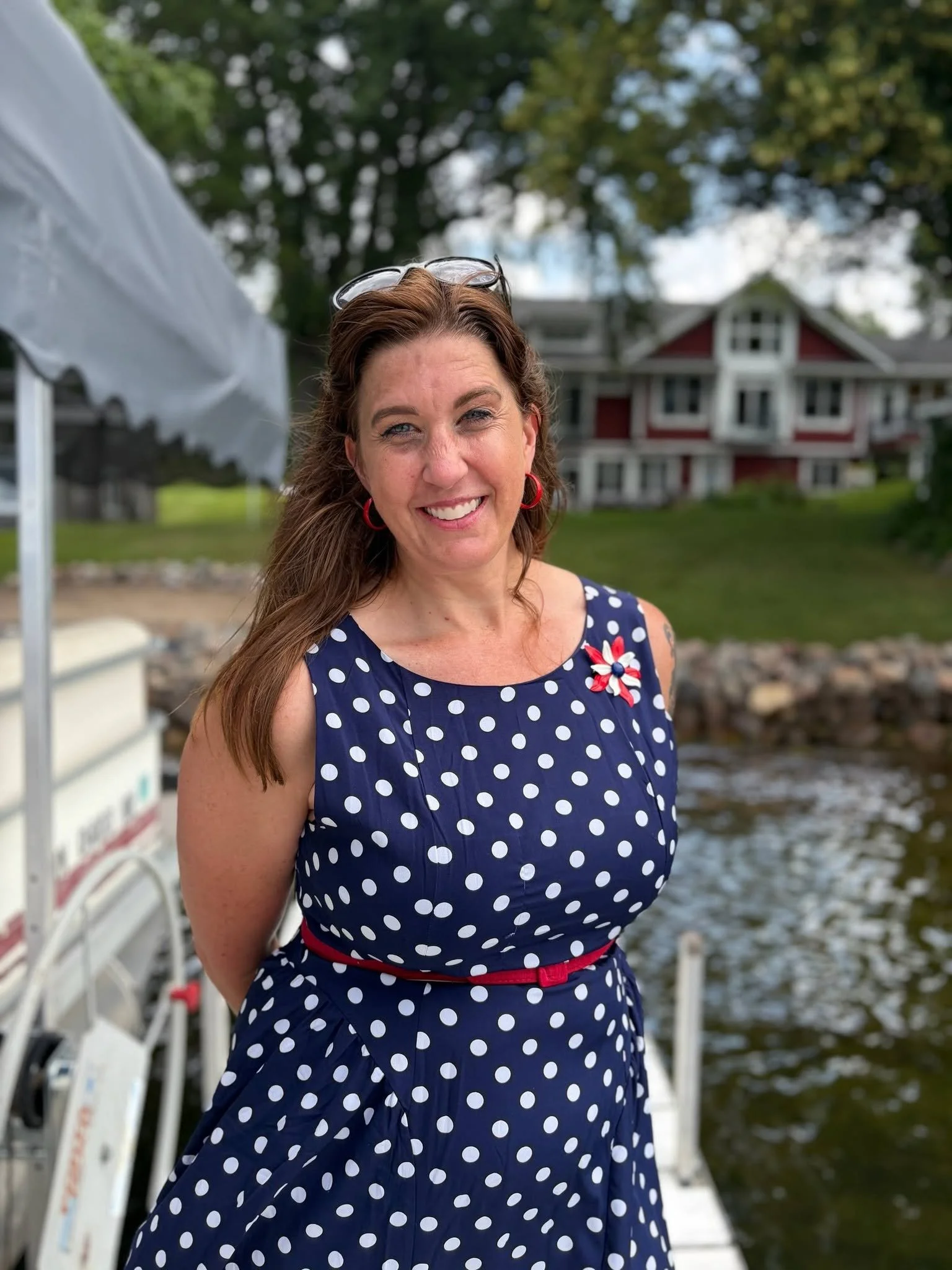 A woman with long brown hair, wearing a navy blue dress with white polka dots and a red belt, smiling outdoors near water, with a house and trees in the background.