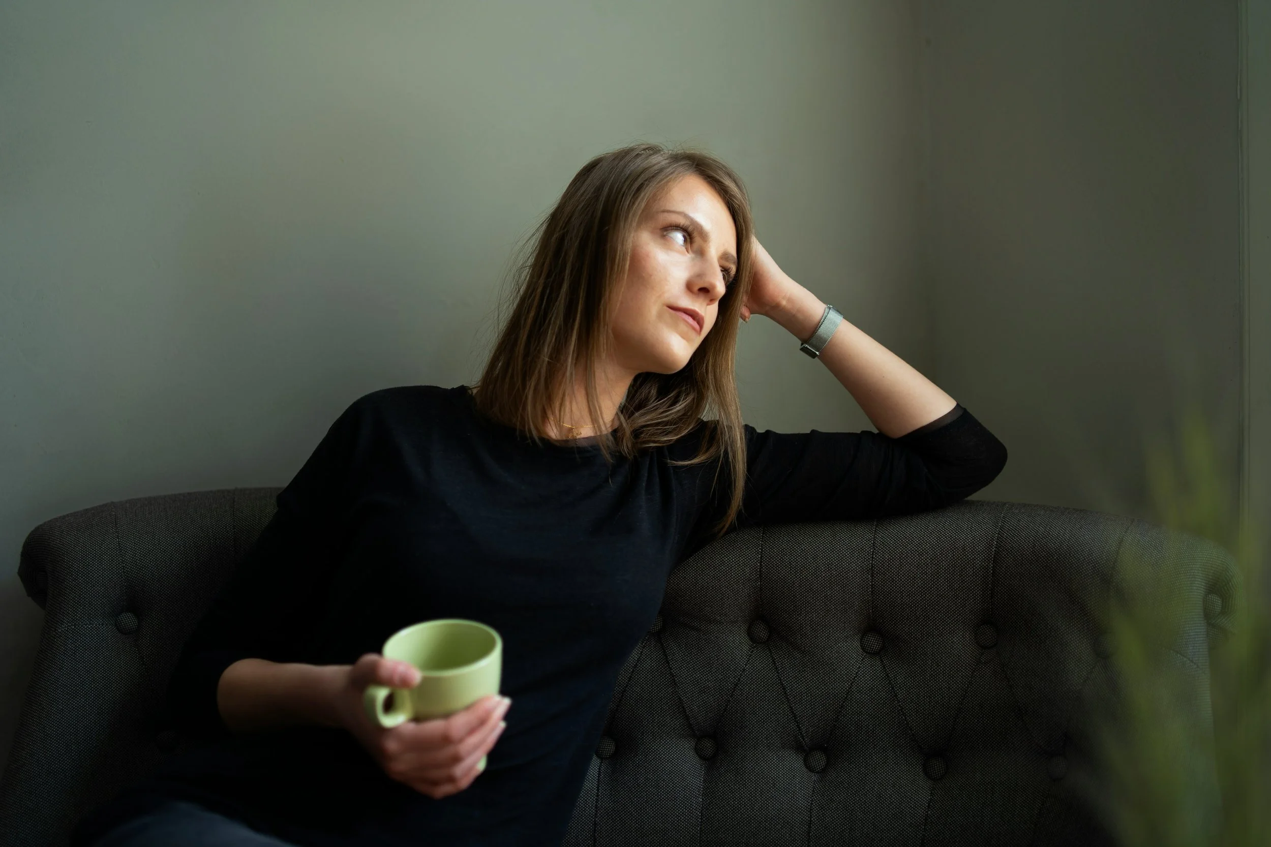 A woman with brown hair wearing a black top is sitting on a dark gray upholstered couch, holding a green mug in her right hand, and looking thoughtfully to her left against a plain light green wall.