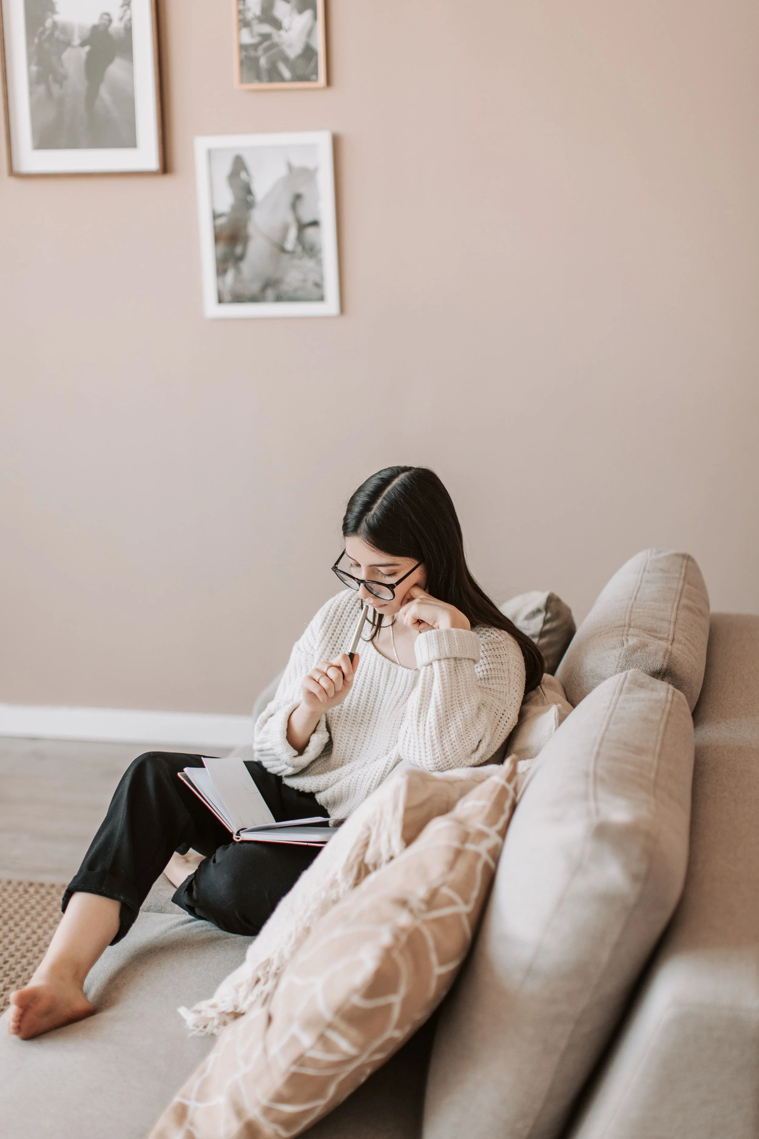 A woman sitting on a beige couch, reading a book with a pen in her hand, in a living room. She is wearing glasses, a white sweater, and black pants. There are pillows on the couch and framed photographs on the wall behind her.