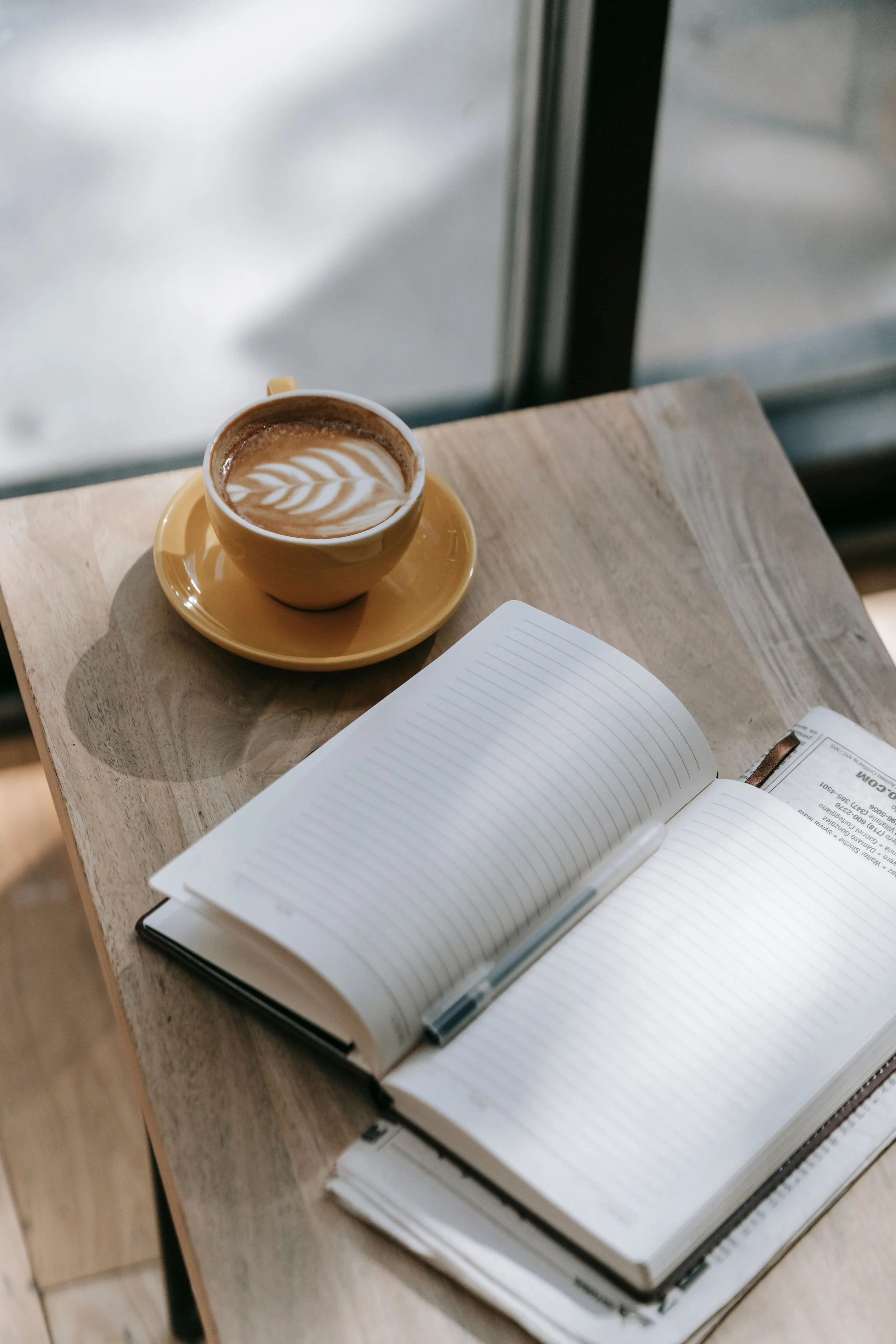 A cup of coffee with latte art on a saucer, placed on a light wooden table, with an open notebook and a pen nearby.