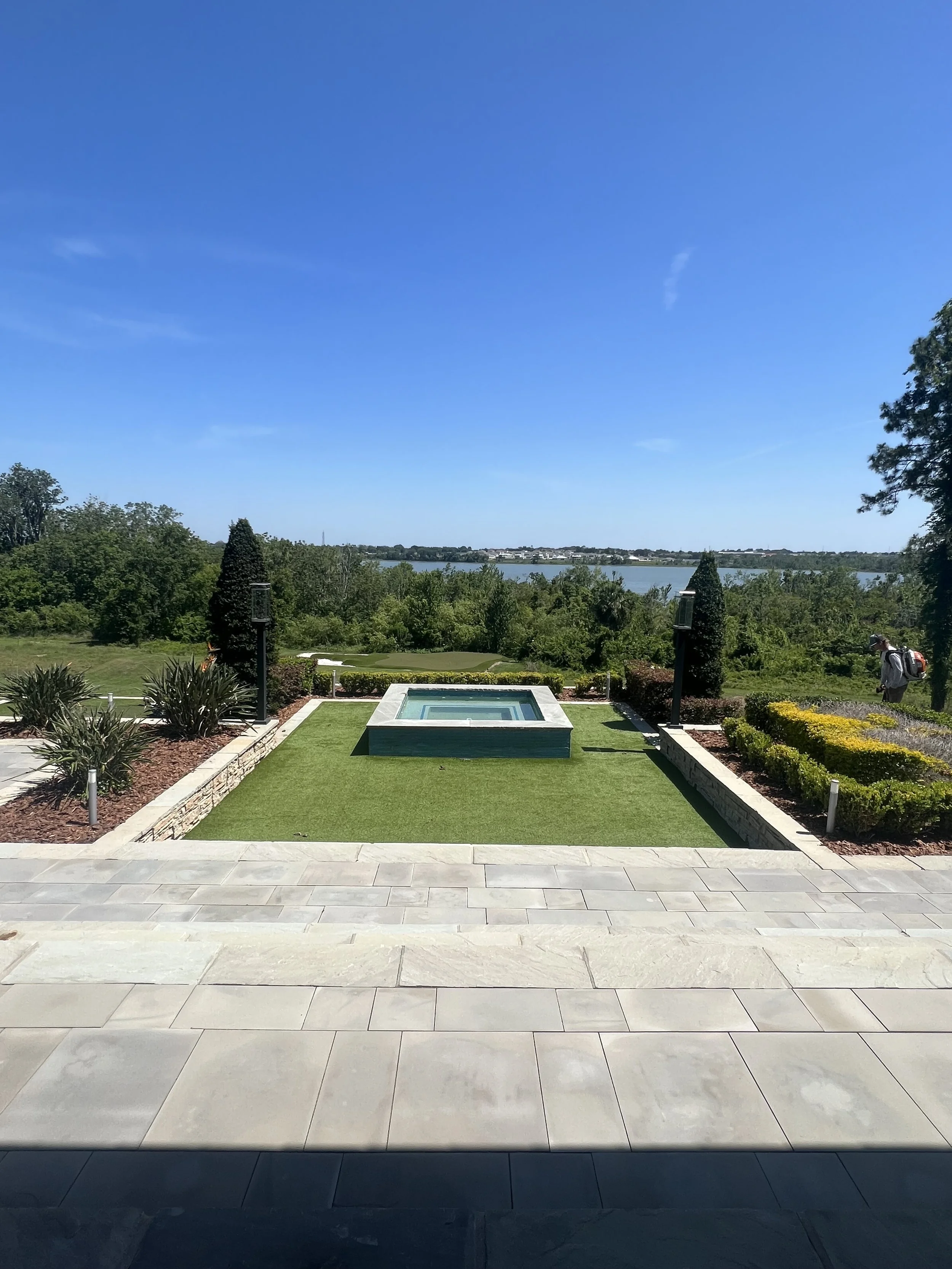 A view from a tiled patio looking out over a manicured garden with a small fountain, lush green trees, a river, and a clear blue sky in the background.