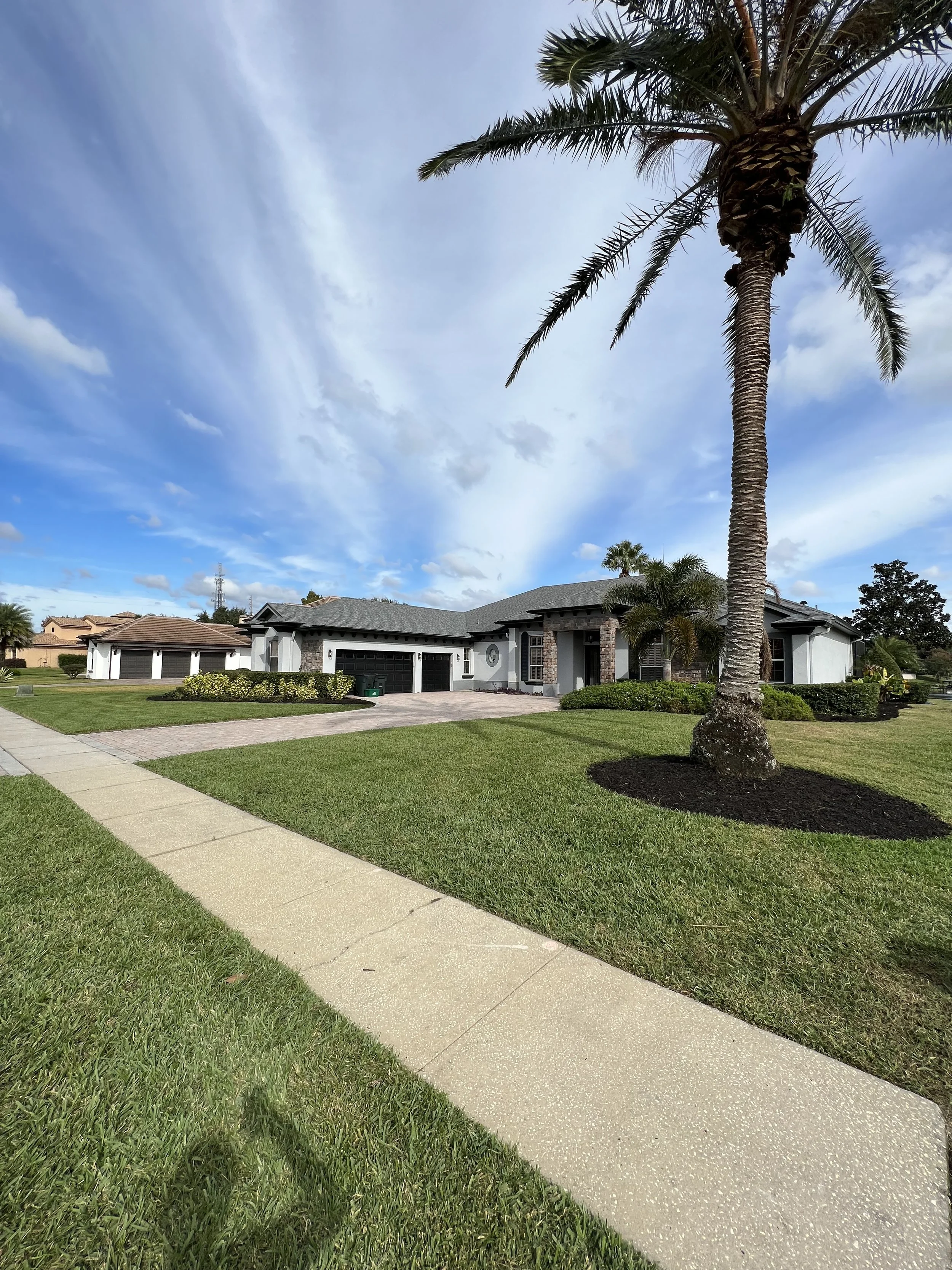 A residential house with a front yard, palm trees, a gravel driveway, and a sidewalk under a partly cloudy sky.