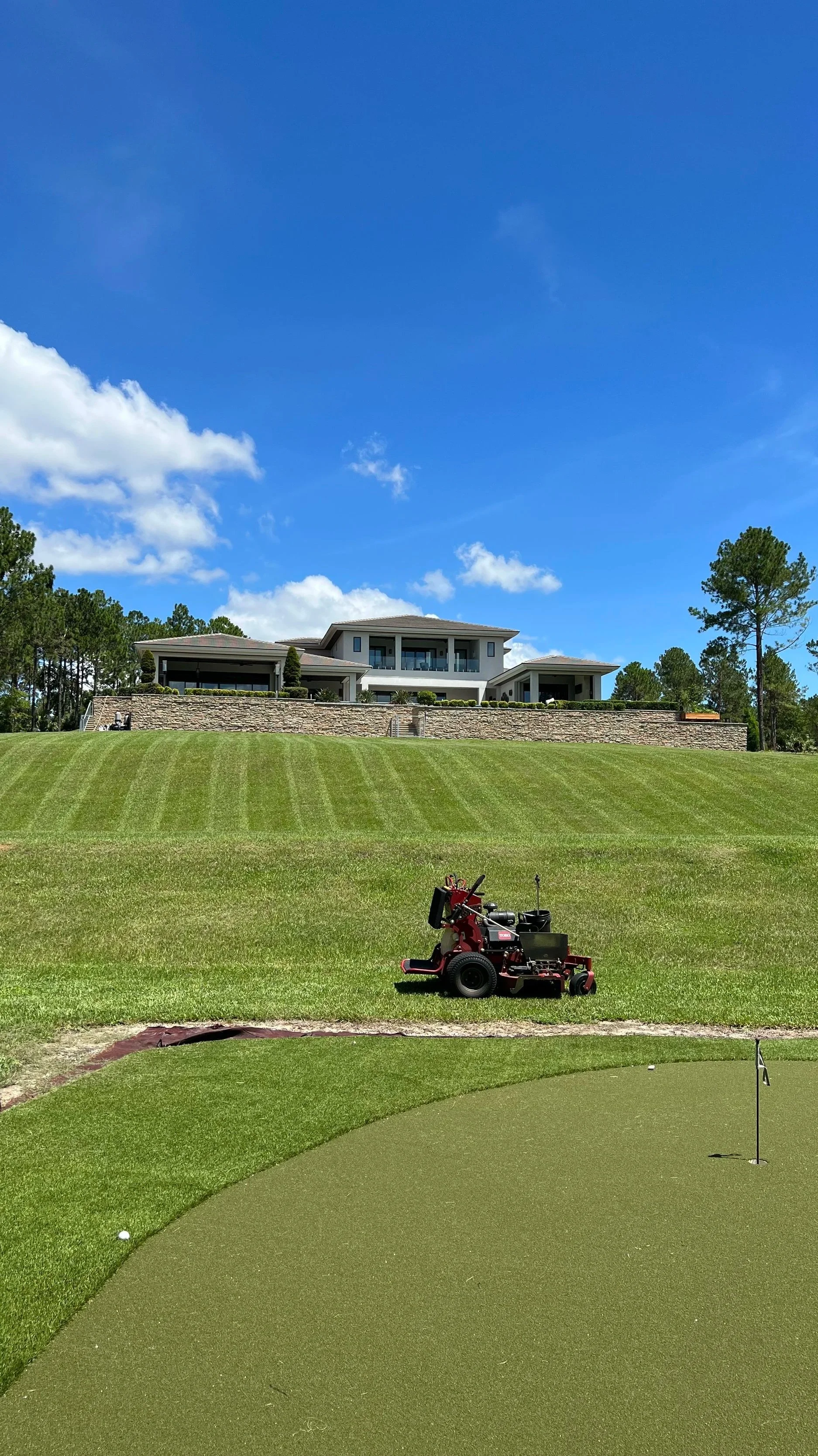 A golf course with a putting green and a golf ball near the hole, a golf cart, and a lawn mower, with a large house and trees in the background under a bright blue sky.
