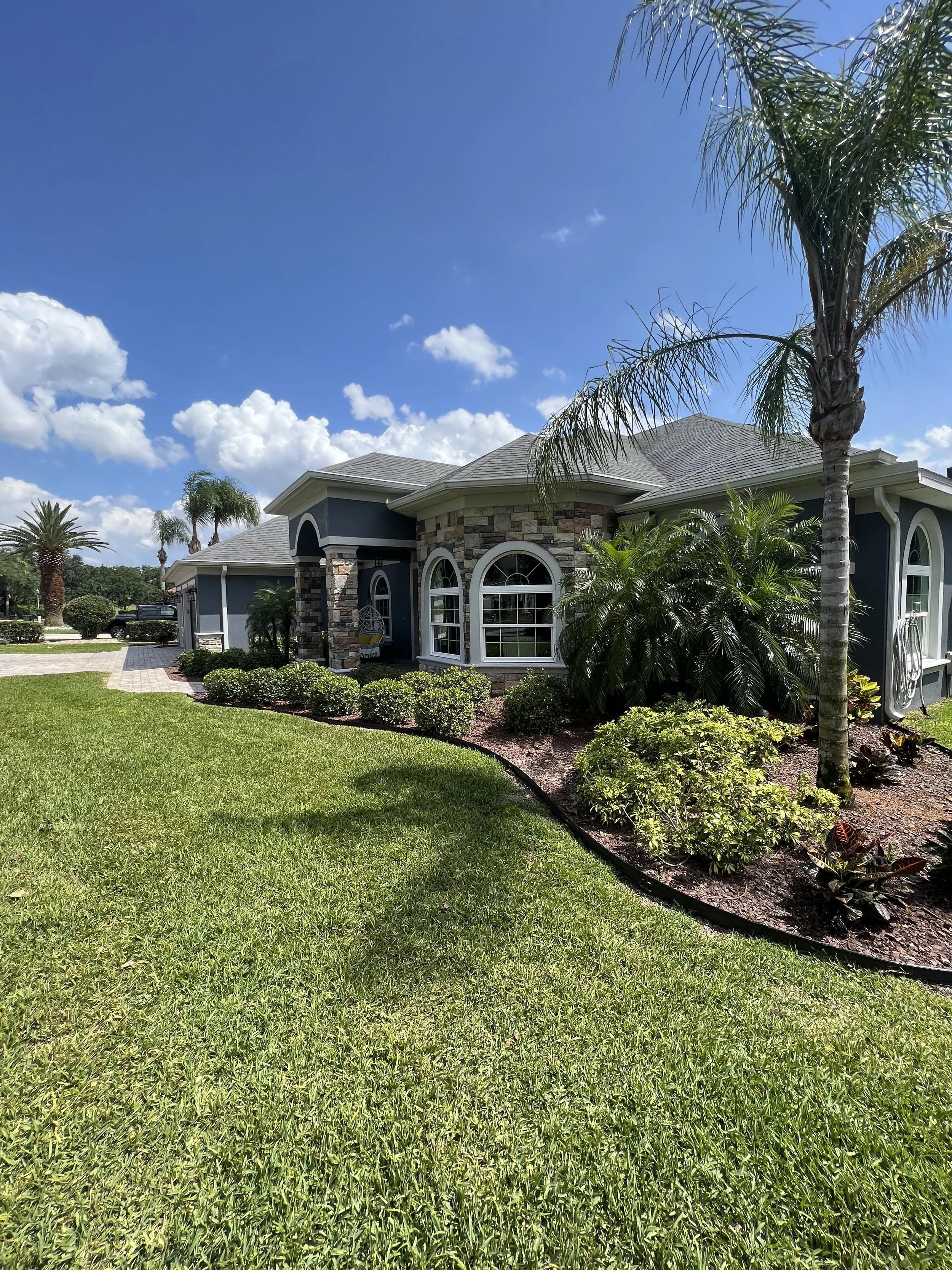 A blue house with stone accents and white trim, surrounded by lush green landscaping and palm trees under a bright blue sky with scattered clouds.
