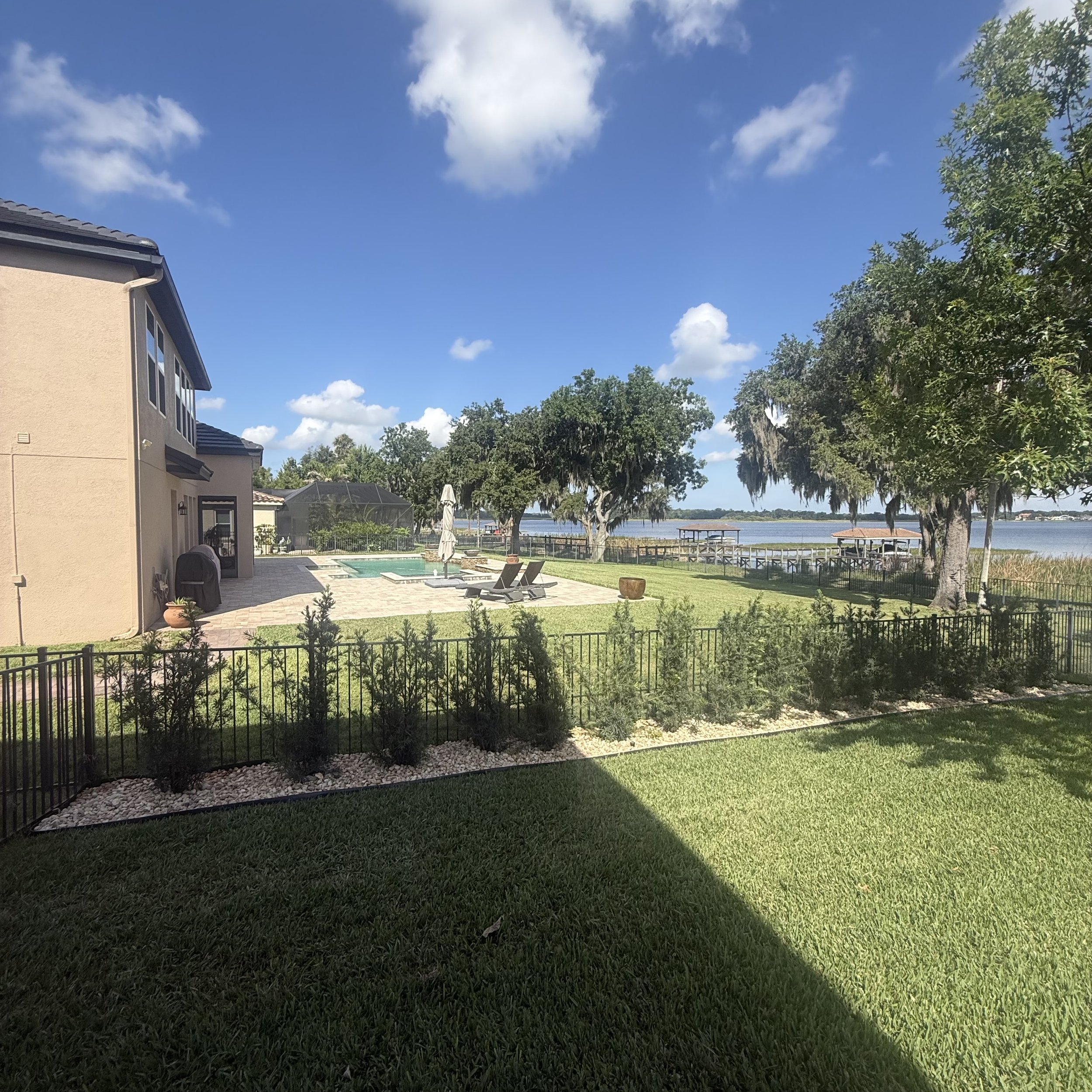 A backyard view overlooking a lake with a patio, lounge chairs, a small pool, surrounding trees, and a house to the left under a partly cloudy blue sky.