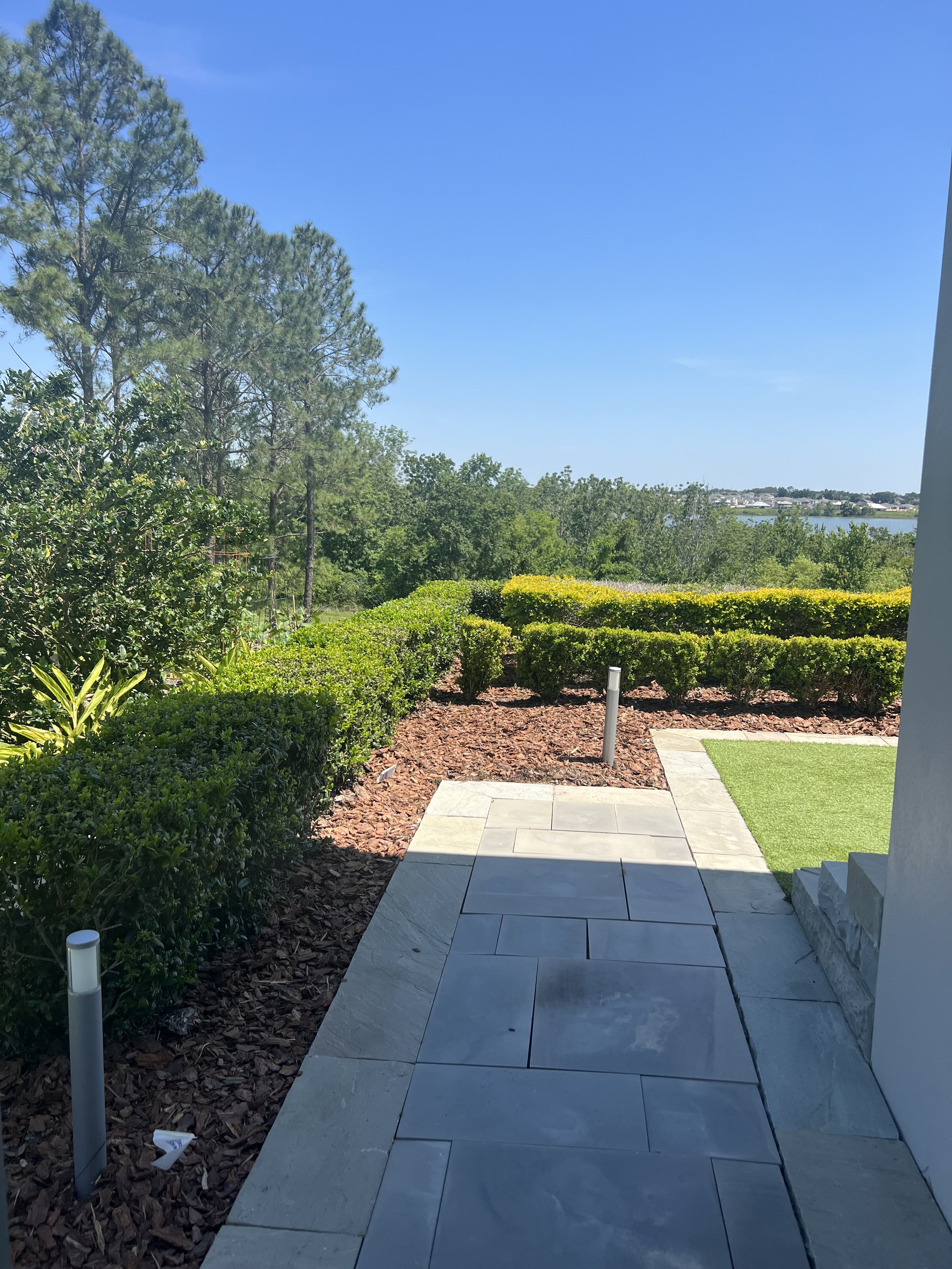 A backyard patio with stone tiles, green shrubs, and a view of trees and a body of water in the distance, under a clear blue sky.